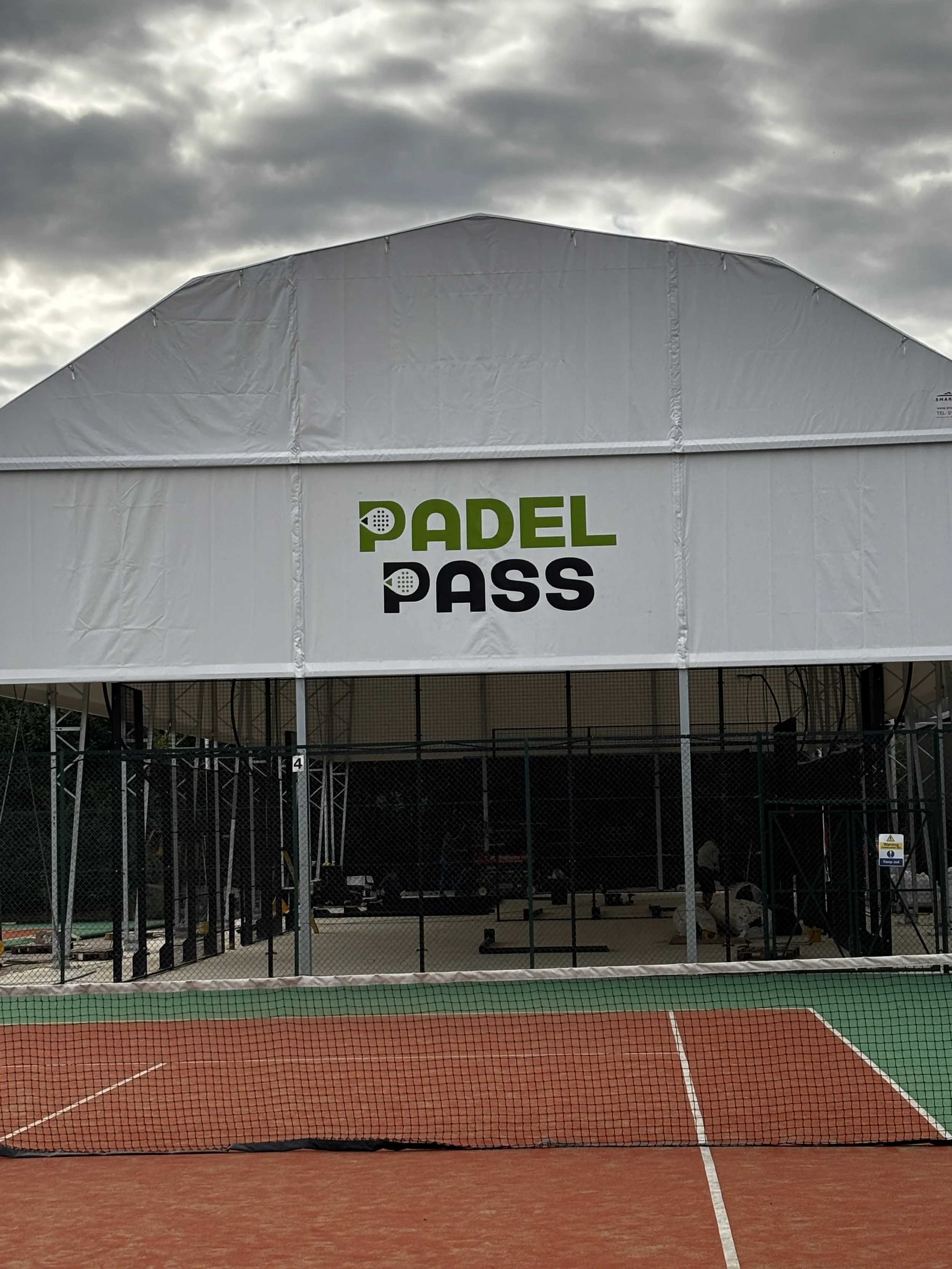 A tennis court with an overhead tent structure displaying the logo 'Padel Pass', with cloudy sky in the background.