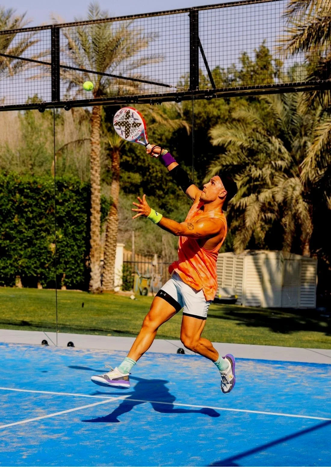 A man playing pickleball on an outdoor court, preparing to hit a ball with a paddle. Palm trees and a fence are visible in the background.