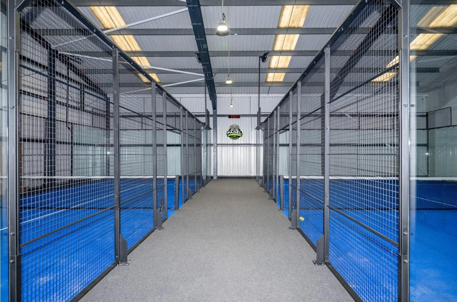 Empty indoor padel tennis courts with metal fencing and blue flooring, seen from the entrance hallway.