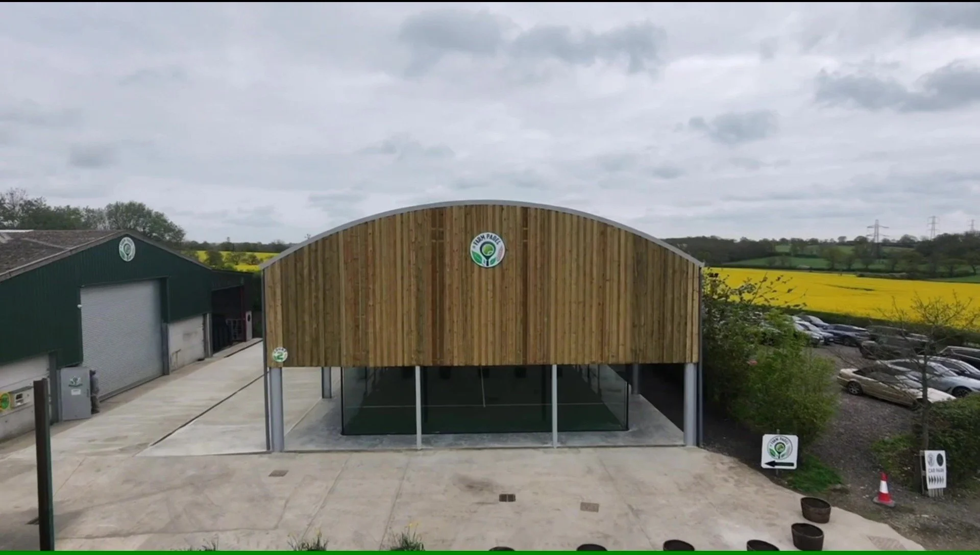 A modern building with a curved wooden facade and a logo reading 'Farm Padel' on the front. There are parking spaces filled with cars and a large green warehouse to the left. Rolling hills, fields, and cloudy skies are visible in the background.