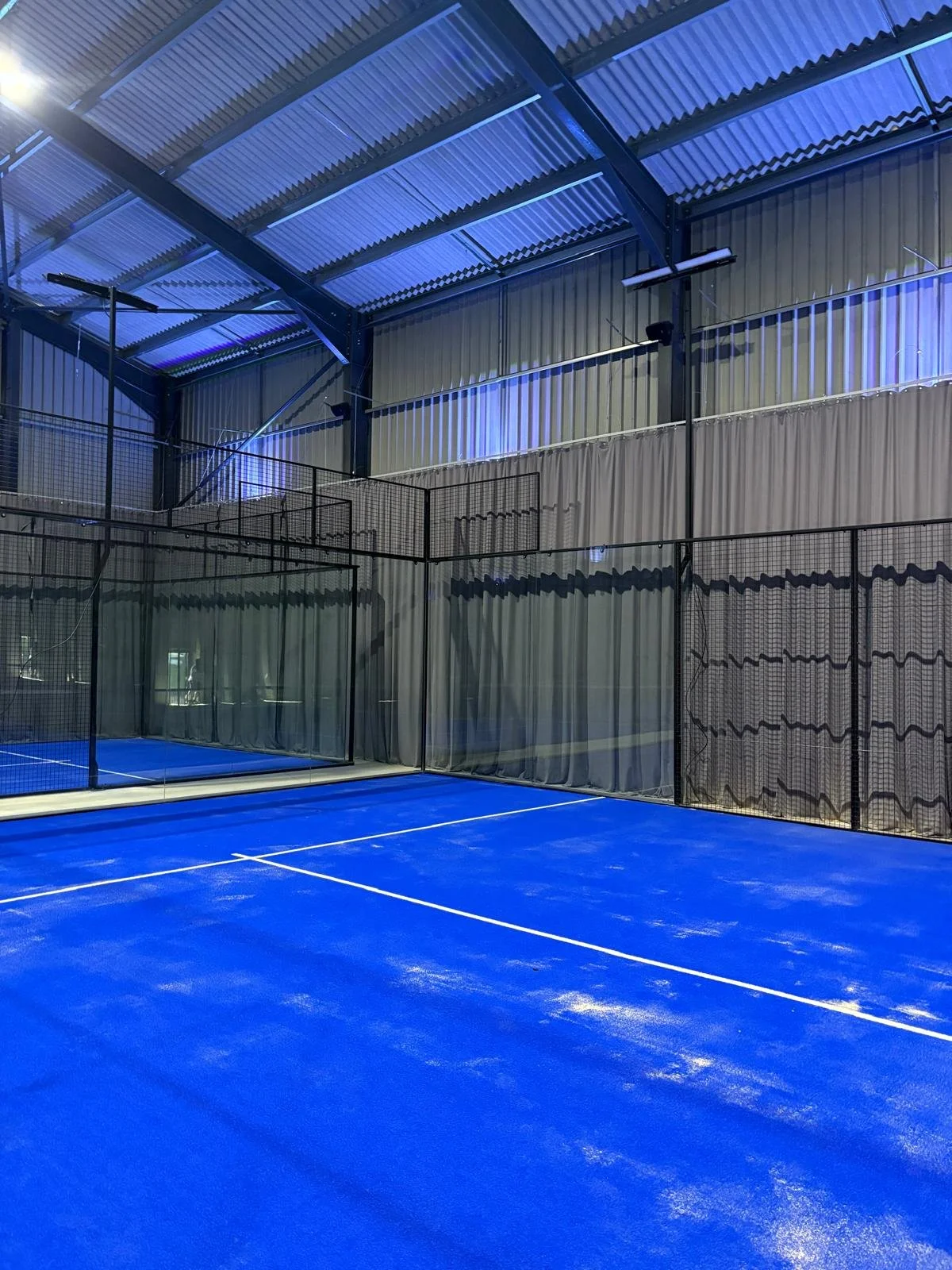 Indoor Padel court with blue surface, surrounded by black netting and grey curtains, with high metal ceiling and blue lighting. 3 Courts in Epping and Ongar
