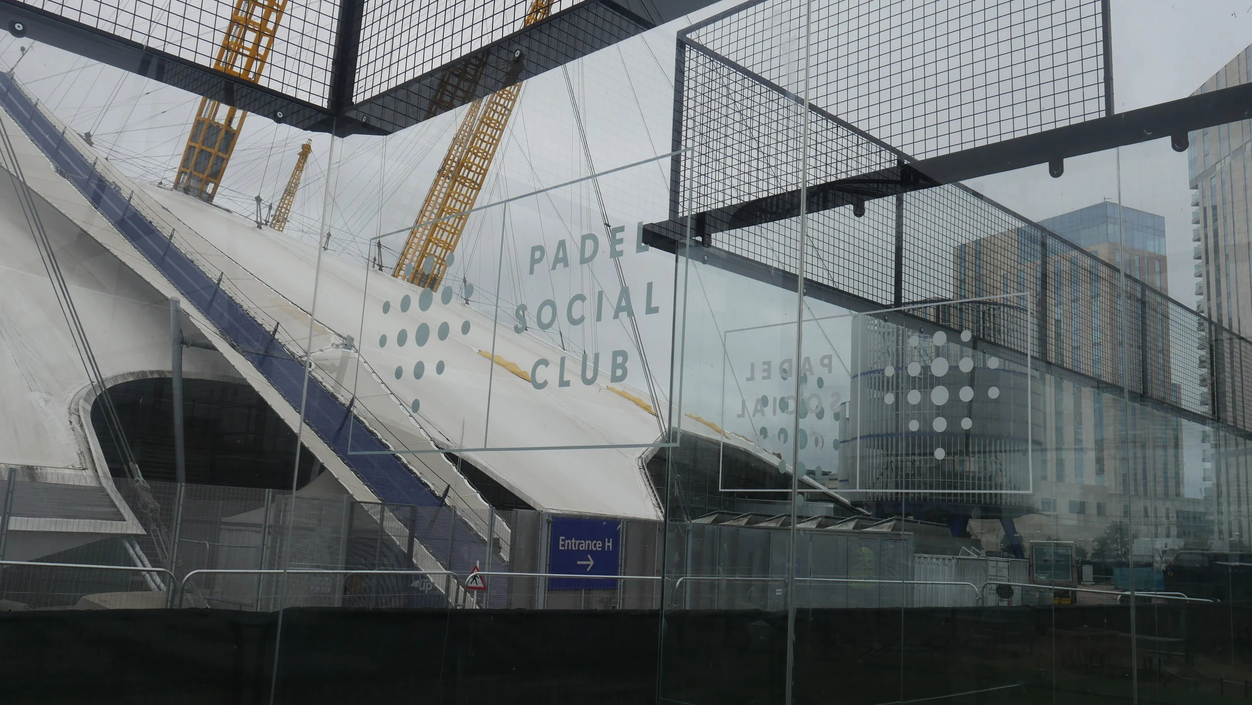 View of a modern sports complex or arena with a steel and glass exterior, featuring signage for a social padel club, with high-rise buildings in the background.