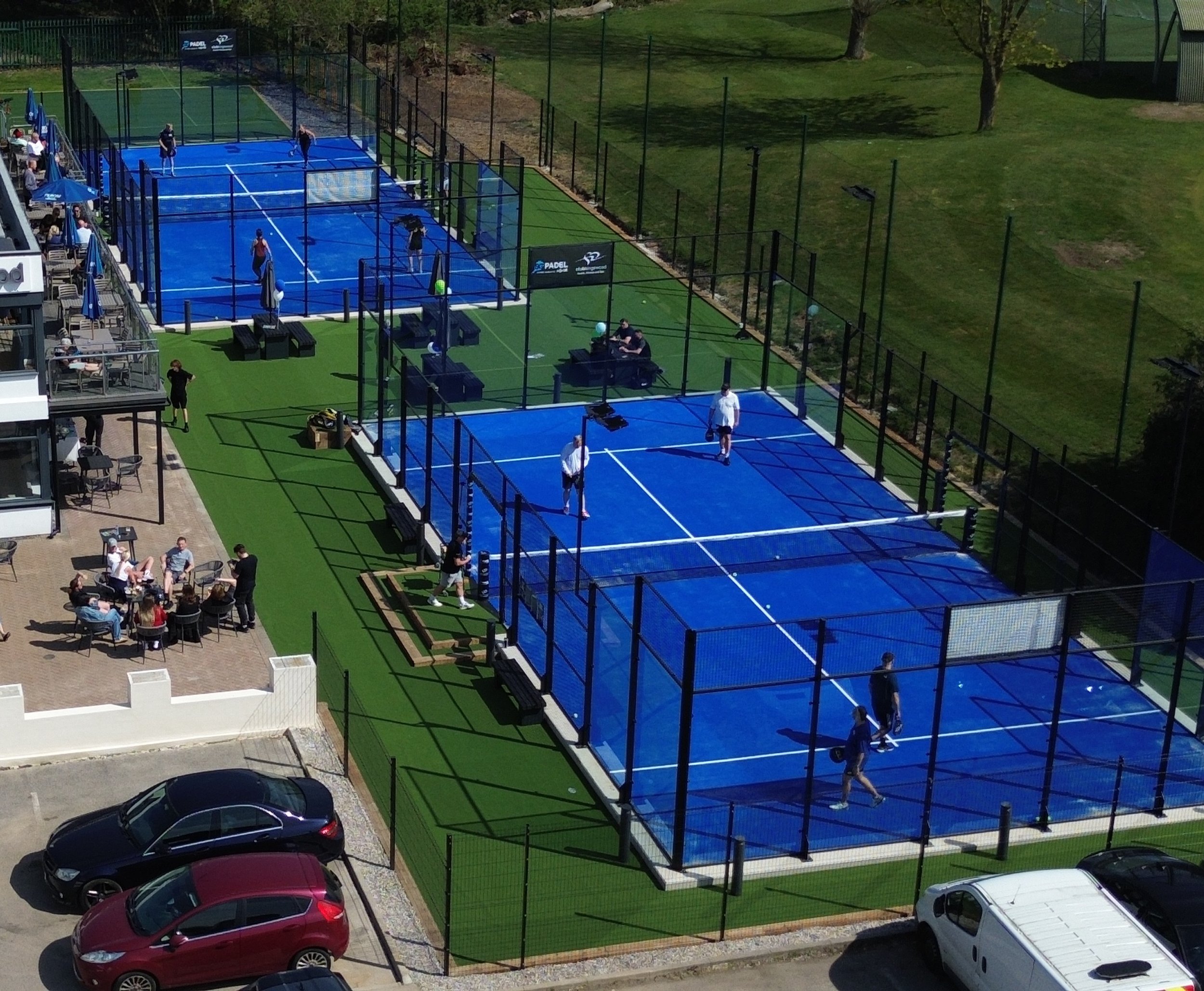 Multiple blue paddle tennis courts with players playing, a seating area with people, parked cars, and a grassy area with trees.