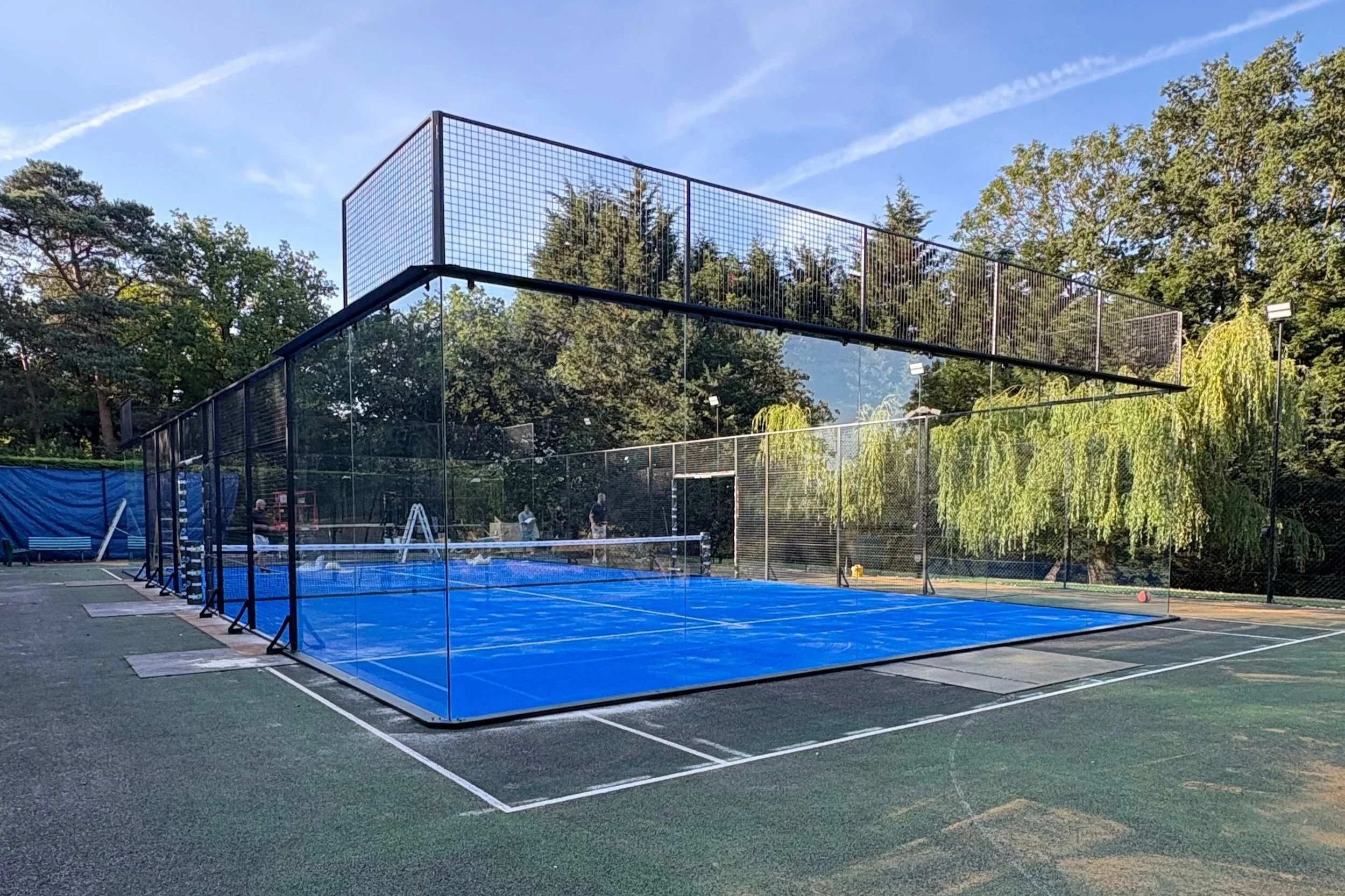 A  Padel tennis court with a blue surface enclosed by a black metal fence, surrounded by trees under a clear blue sky.