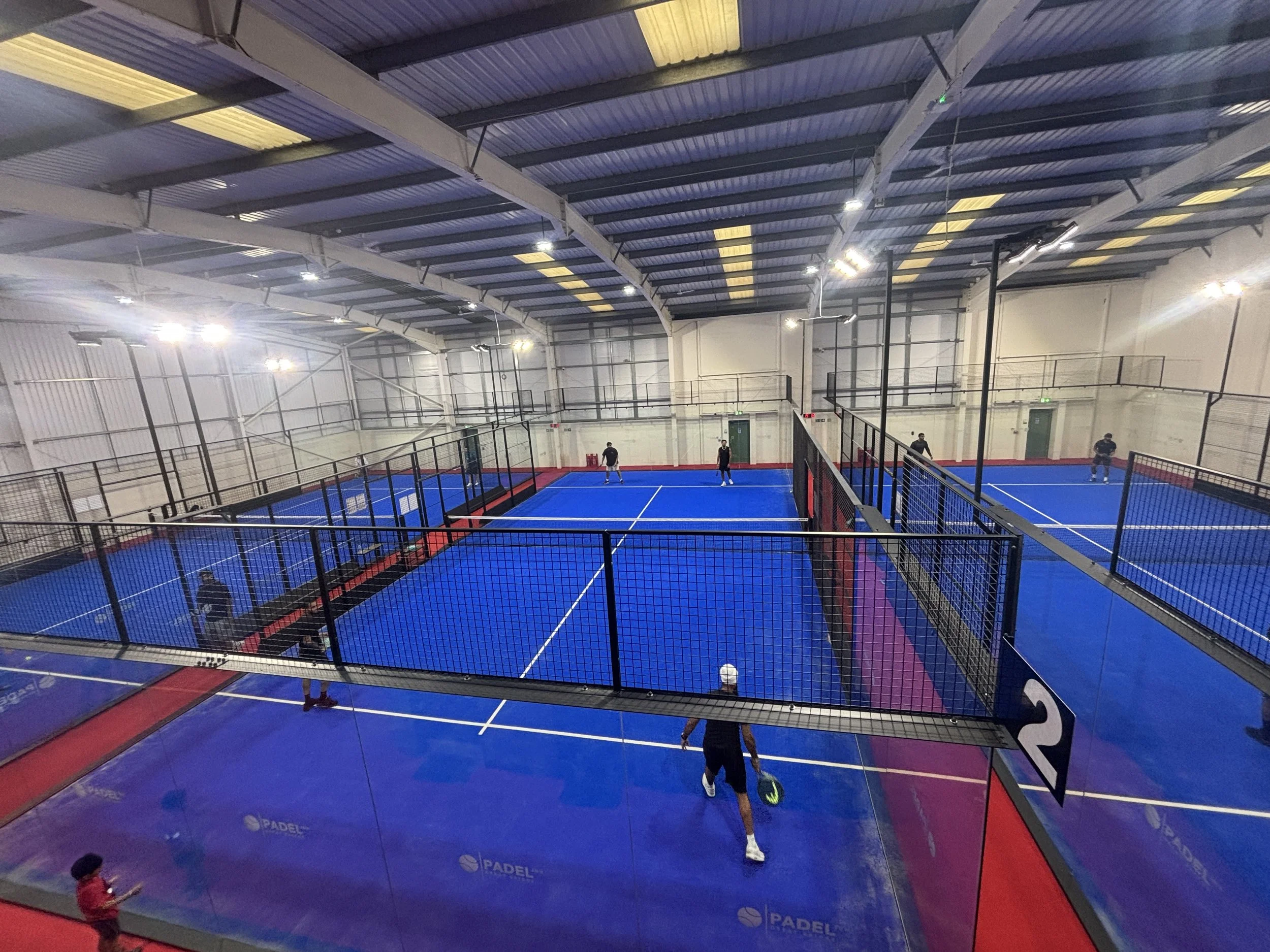 Indoor padel tennis court with blue playing surface surrounded by glass walls and black fencing UKs Best Padel