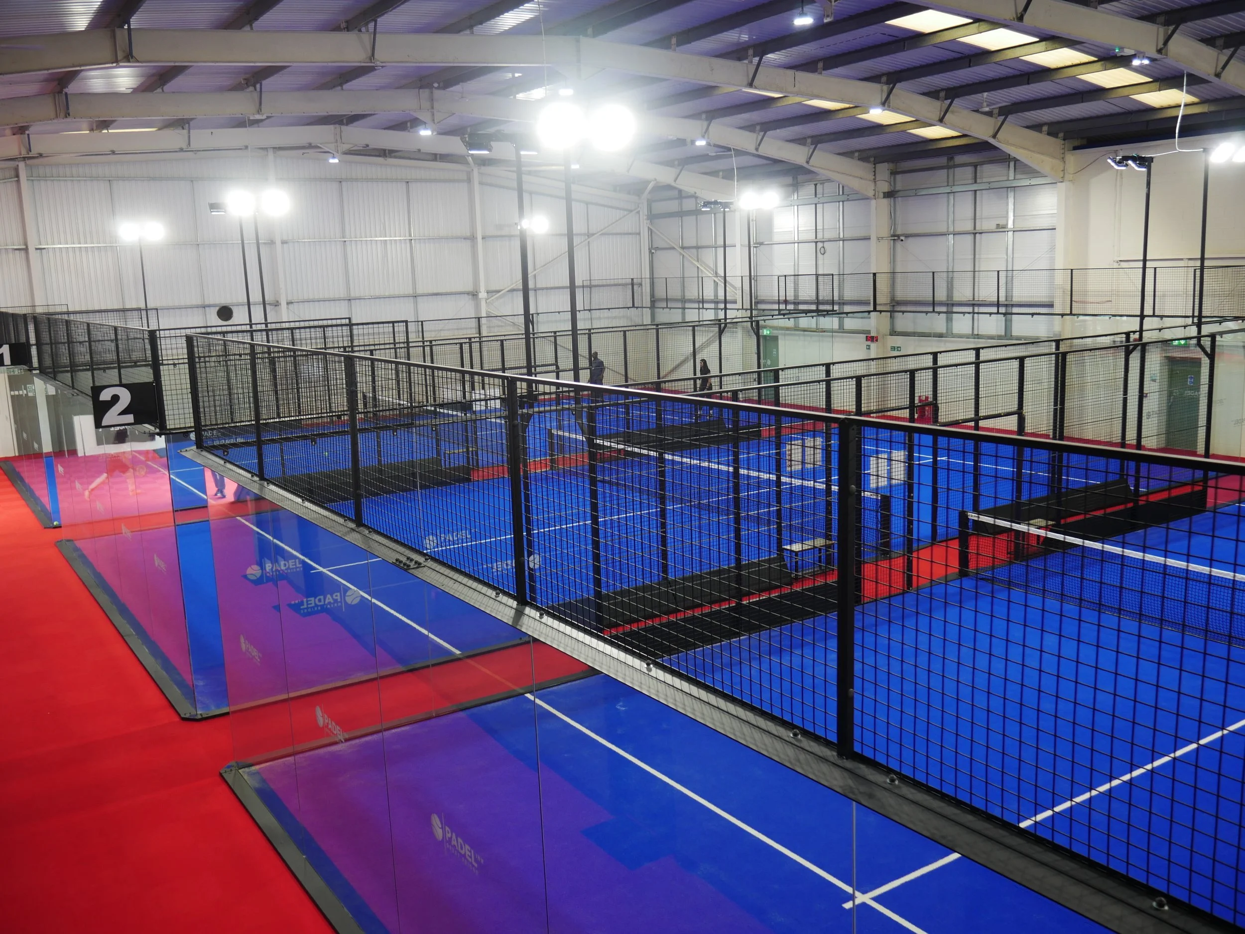 Indoor padel courts with blue surfaces, black fencing, and a red perimeter, inside a large warehouse-style building in the UK