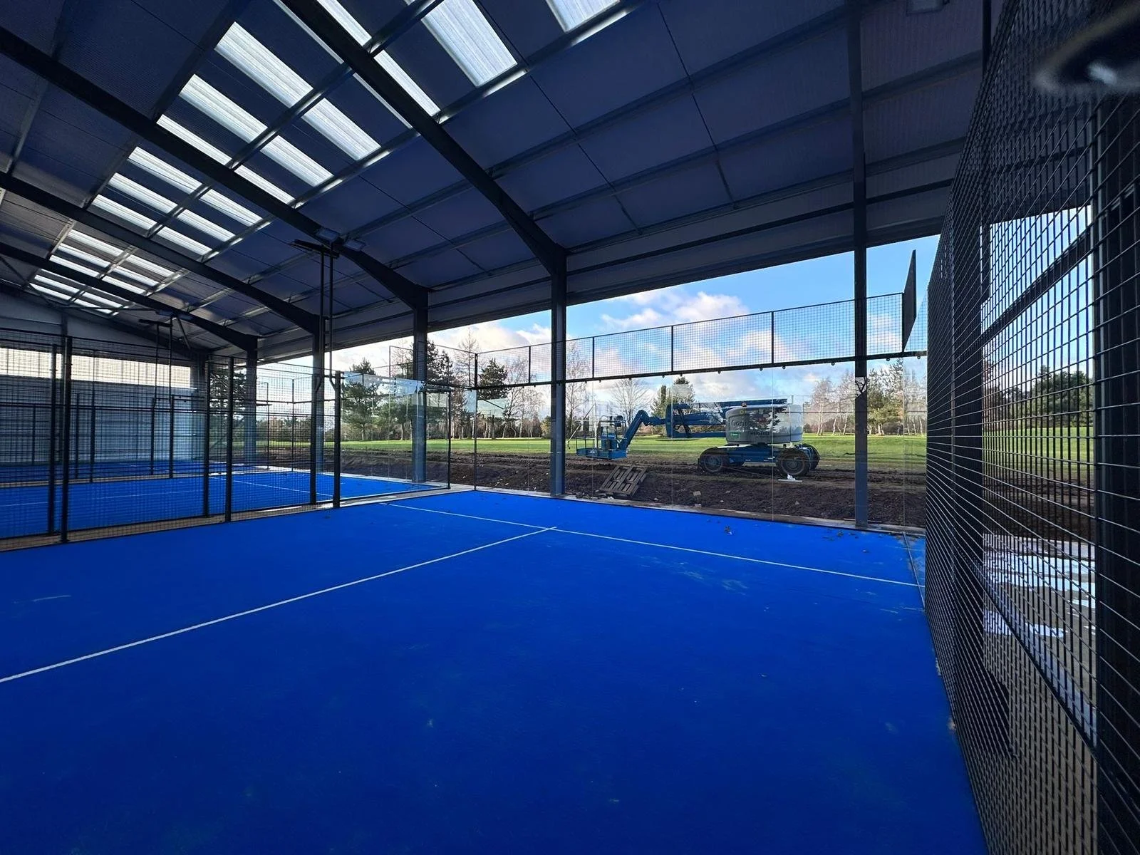  padel court with a bright blue surface, underneath a large Canopy in the UK surrounded by black mesh fencing, overlooking an outdoor field with construction equipment, trees, and a blue sky outside.