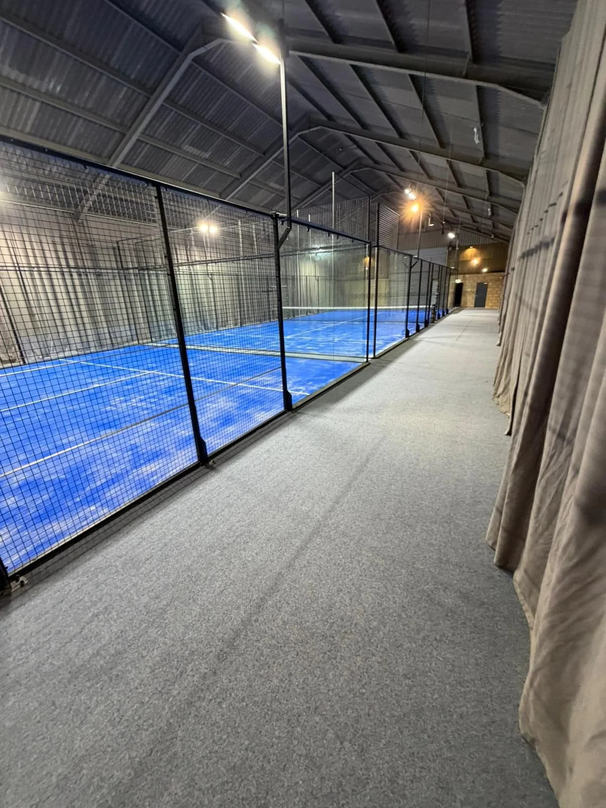 Indoor Mejorset Padel courts with blue surface and grey acoustic curtains, illuminated by ceiling lights at night.