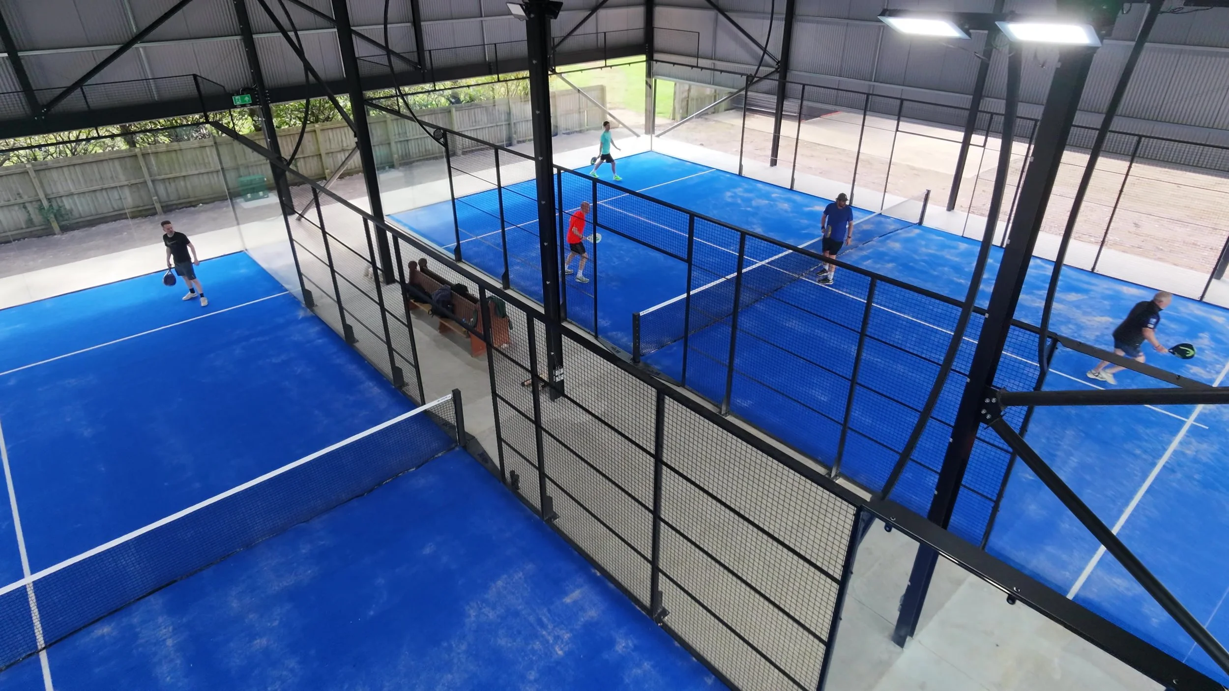 Indoor padel courts with three players playing, two on one court and one on the other, with a seating area and fence surrounding the courts.