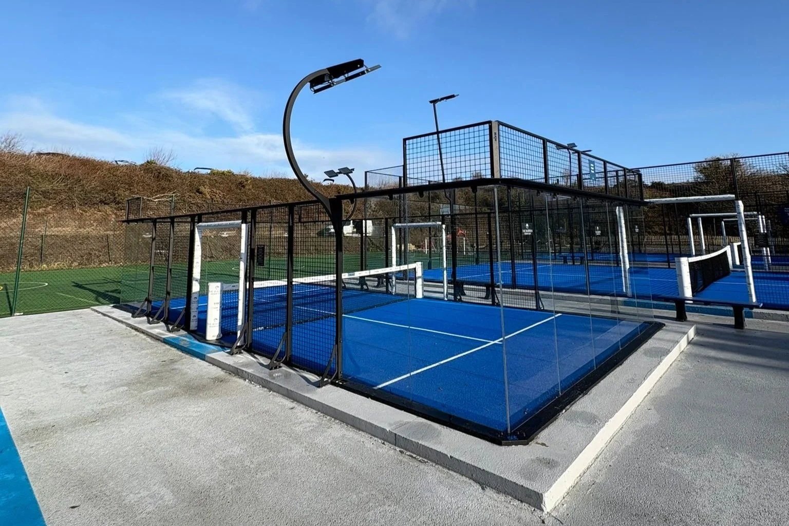 Outdoor sports complex with multiple pickleball courts enclosed by a black metal fence, situated on a concrete surface with a hillside and blue sky in the background.