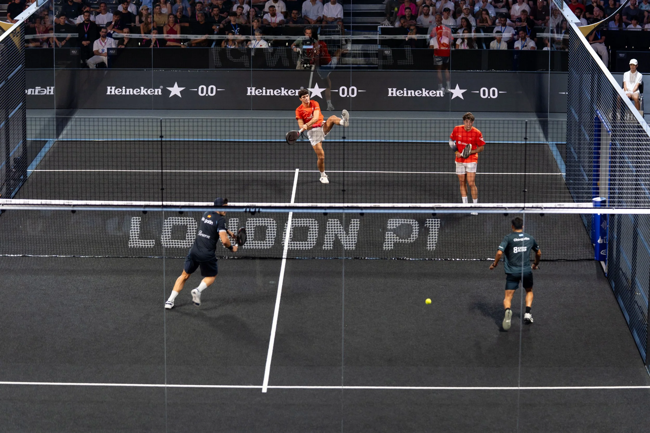 Two male players in red shirts and white shorts playing a game of padel tennis on an outdoor court surrounded by a tall glass fence. One player is in mid-air hitting a ball, while the other is on the court ready for the next move. In the background, spectators are seated in stands watching the match.
