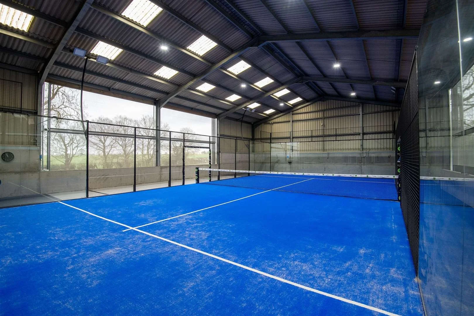 Indoor Padel court with blue surface, net in the middle, enclosed by glass walls and a metal roof with skylights, outside trees are visible through the open side.