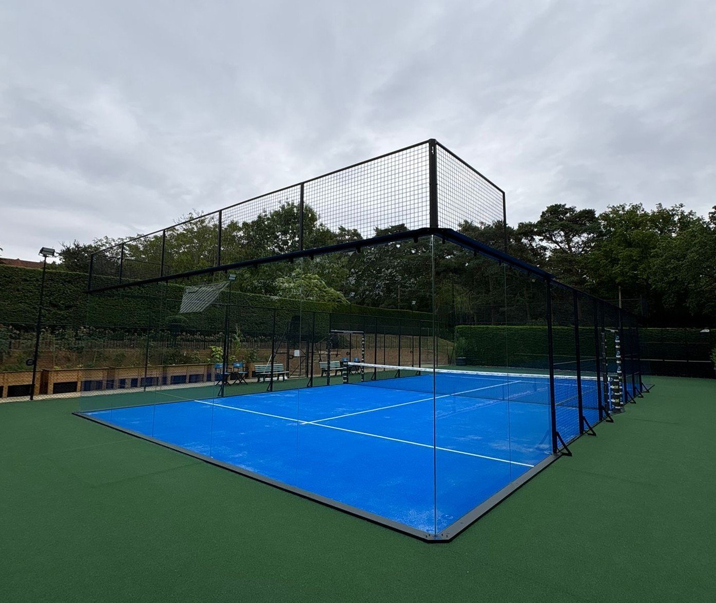 An outdoor padel tennis court with blue surface, Surrounded by green turf.
