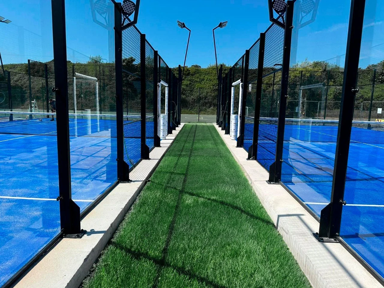 A pickleball court with blue surface, surrounded by black fencing, and a green artificial grass walkway in the middle, under a bright blue sky with some trees in the background.