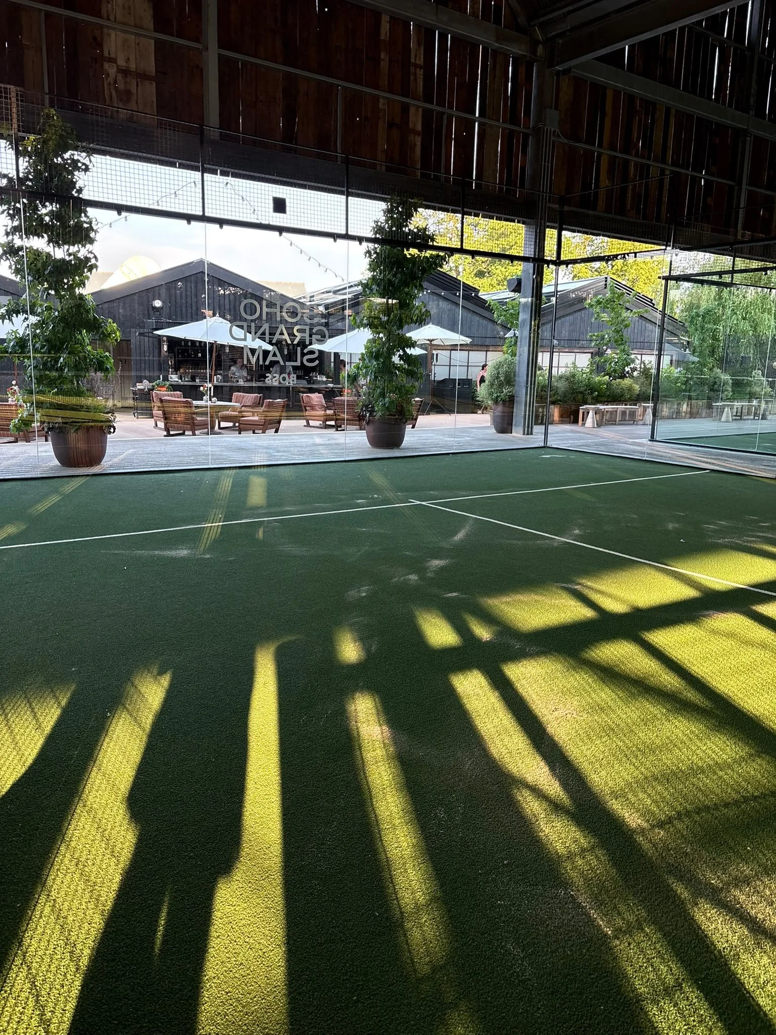 Soho Farmhouse Padel court with a green artificial turf surface, shadows from overhead structure, and a glass wall showing an outdoor seating area with potted plants and umbrellas.