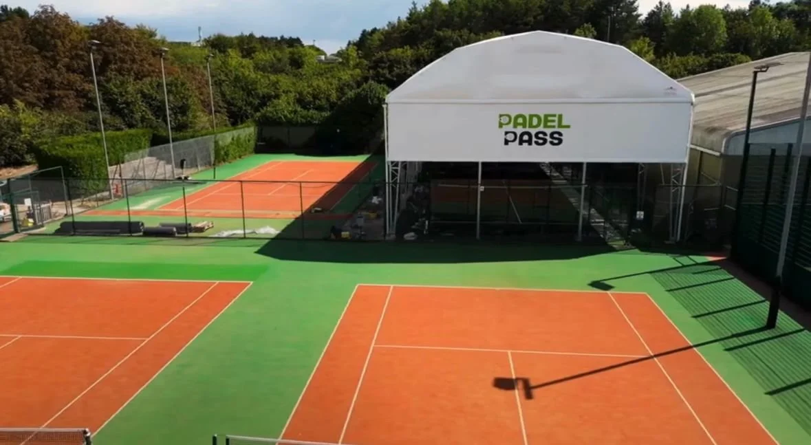 Empty outdoor tennis courts adjacent to a UK Padel Site underneath a Large Canopy with "Padel Pass" written across it , surrounded by green trees and fencing.