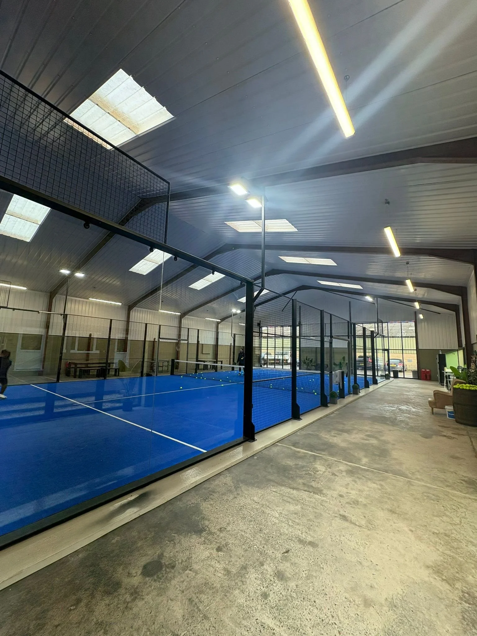 Indoor Padel facility with a blue Padel court, enclosed by black mesh fencing, and illuminated by overhead lights.