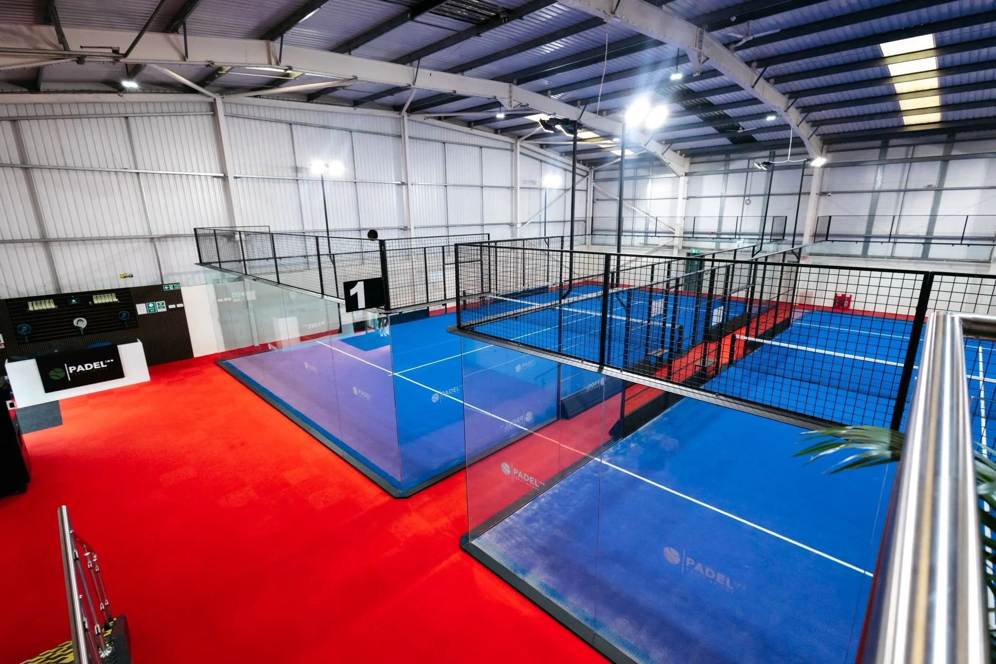 Indoor padel tennis court with blue playing surface surrounded by glass walls and black fencing UKs Best Padel
