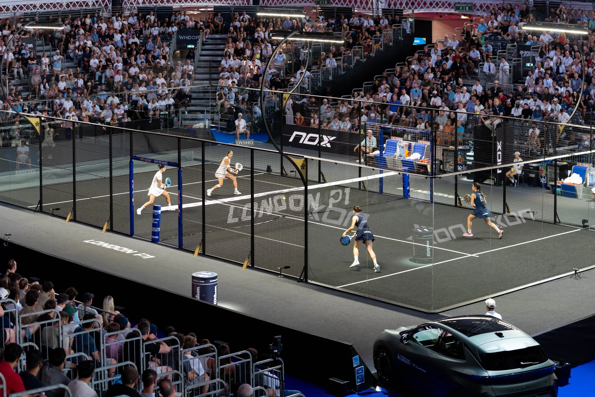 A professional indoor padel tennis match with players on a court enclosed by a glass fence, surrounded by a large audience seated in tiered seating, with some spectators standing. The court is branded and there are chairs and equipment beside the court, indicating a formal tournament setting.