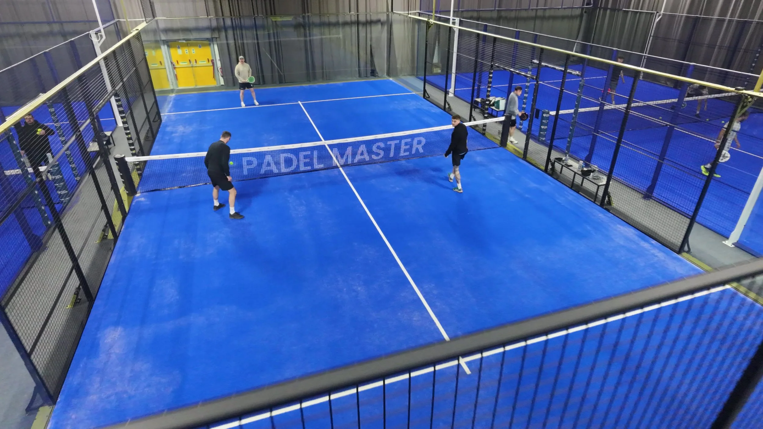 Indoor padel court with blue playing surface, surrounded by black fencing and net, and equipped with multiple players practicing and playing.