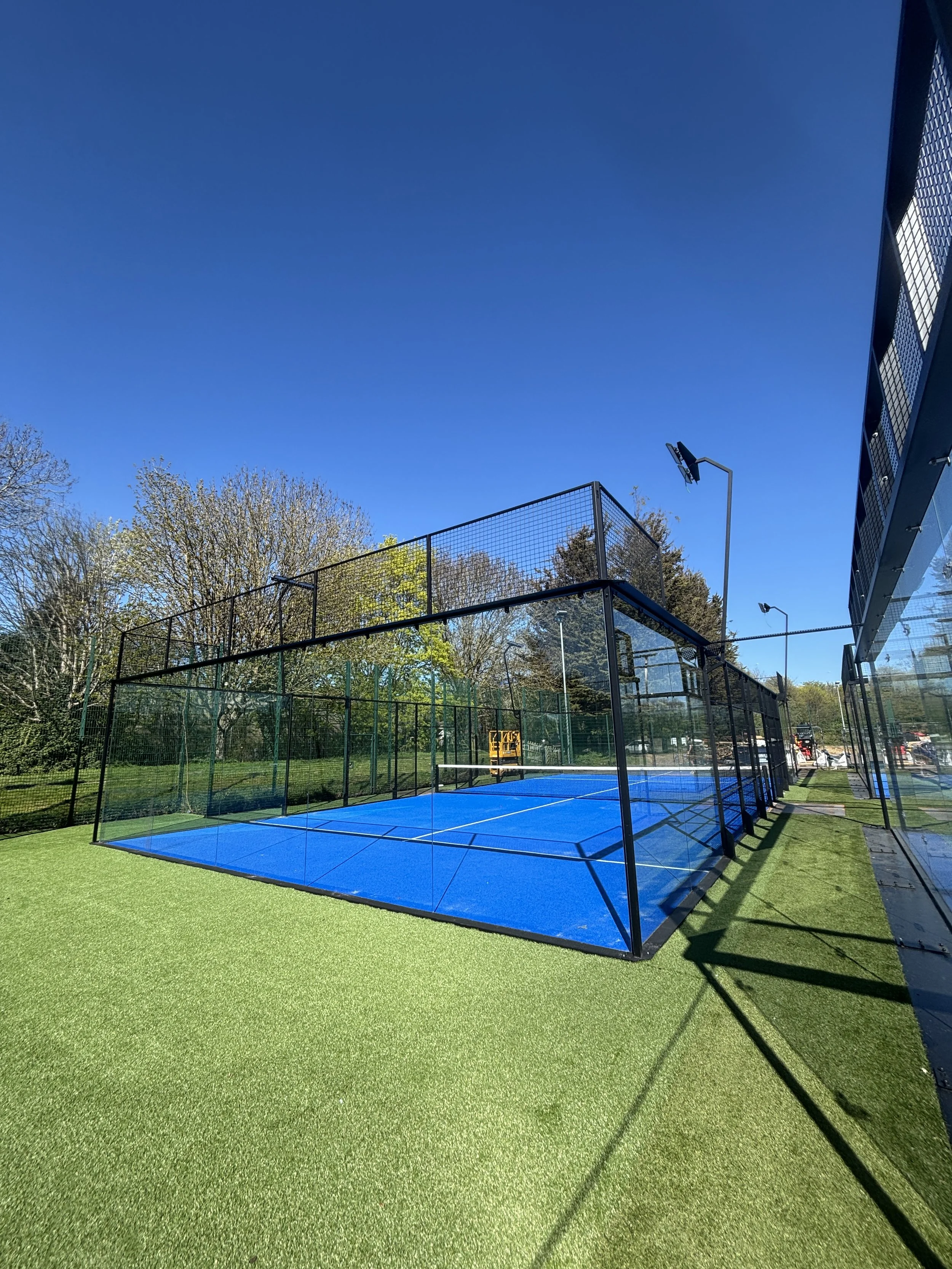 A bright blue Padel court with a netted fence surrounding it, adjacent to green artificial grass and trees, with a clear blue sky overhead.