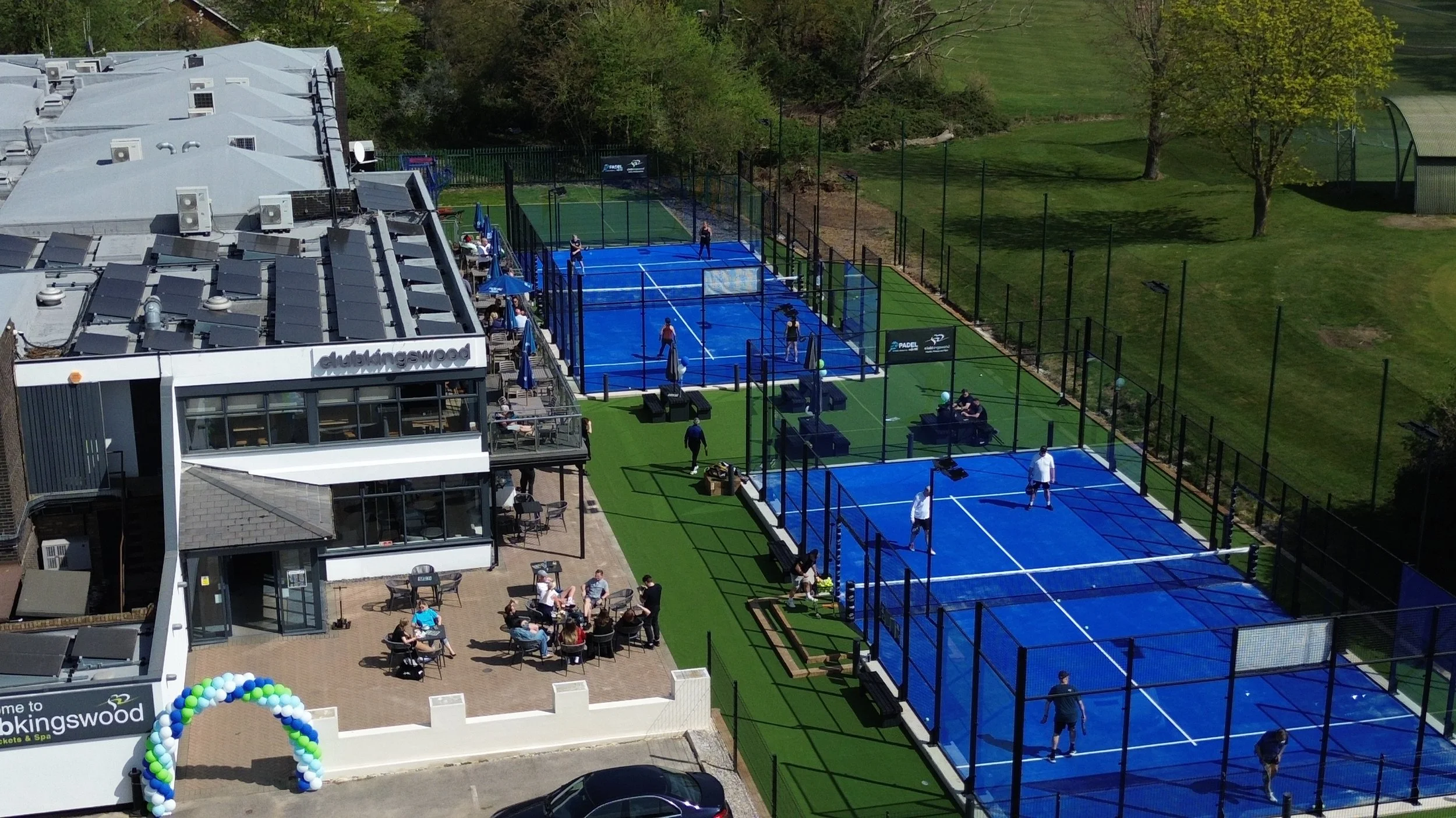 Aerial view of a tennis and padel court complex at Club kingswood with multiple blue courts, people playing, and an outdoor seating area with tables and chairs.