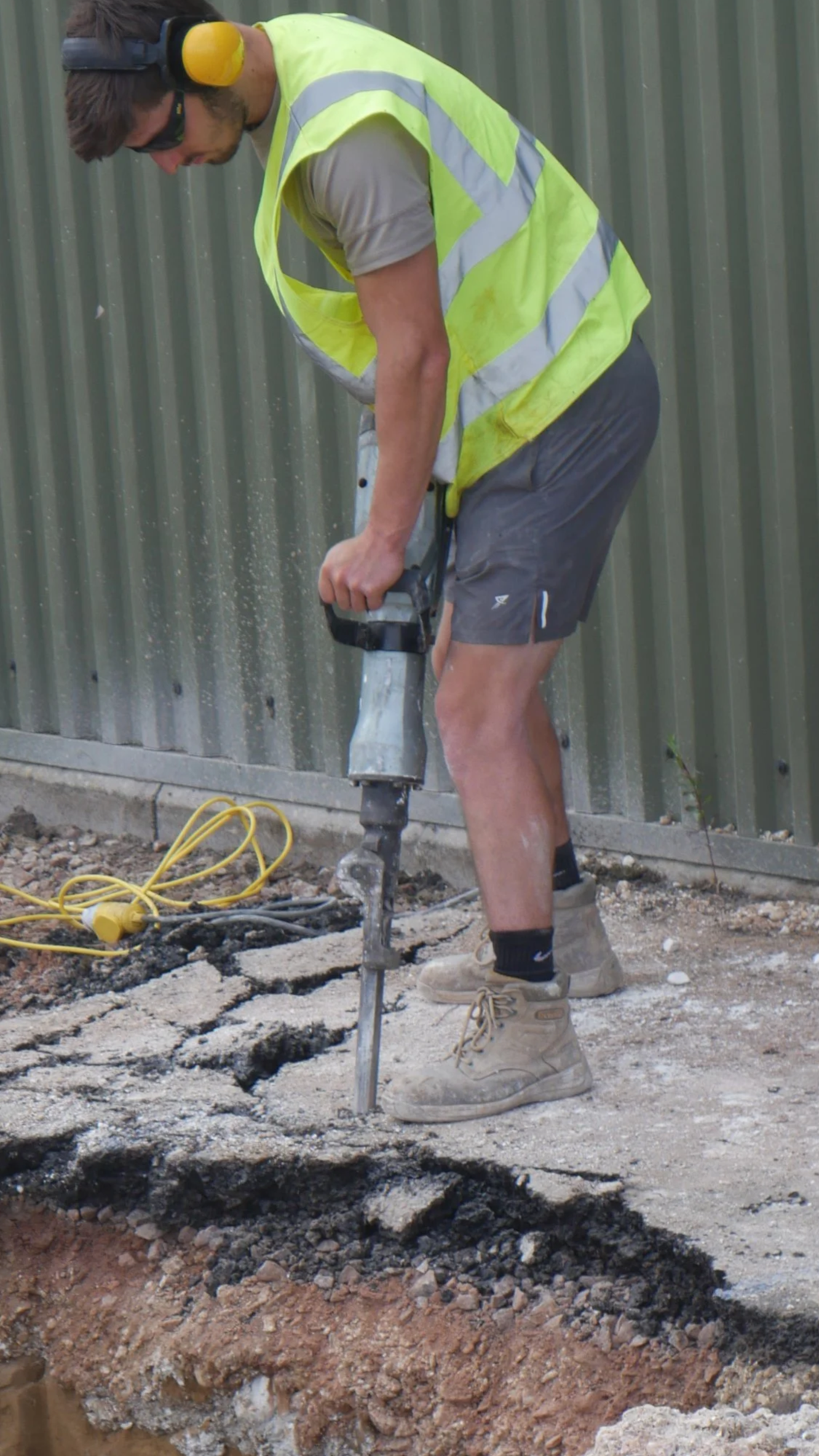A construction worker wearing a yellow safety vest, beige boots, and dark shorts is using a jackhammer to break up pavement on a construction site for Padel Courts