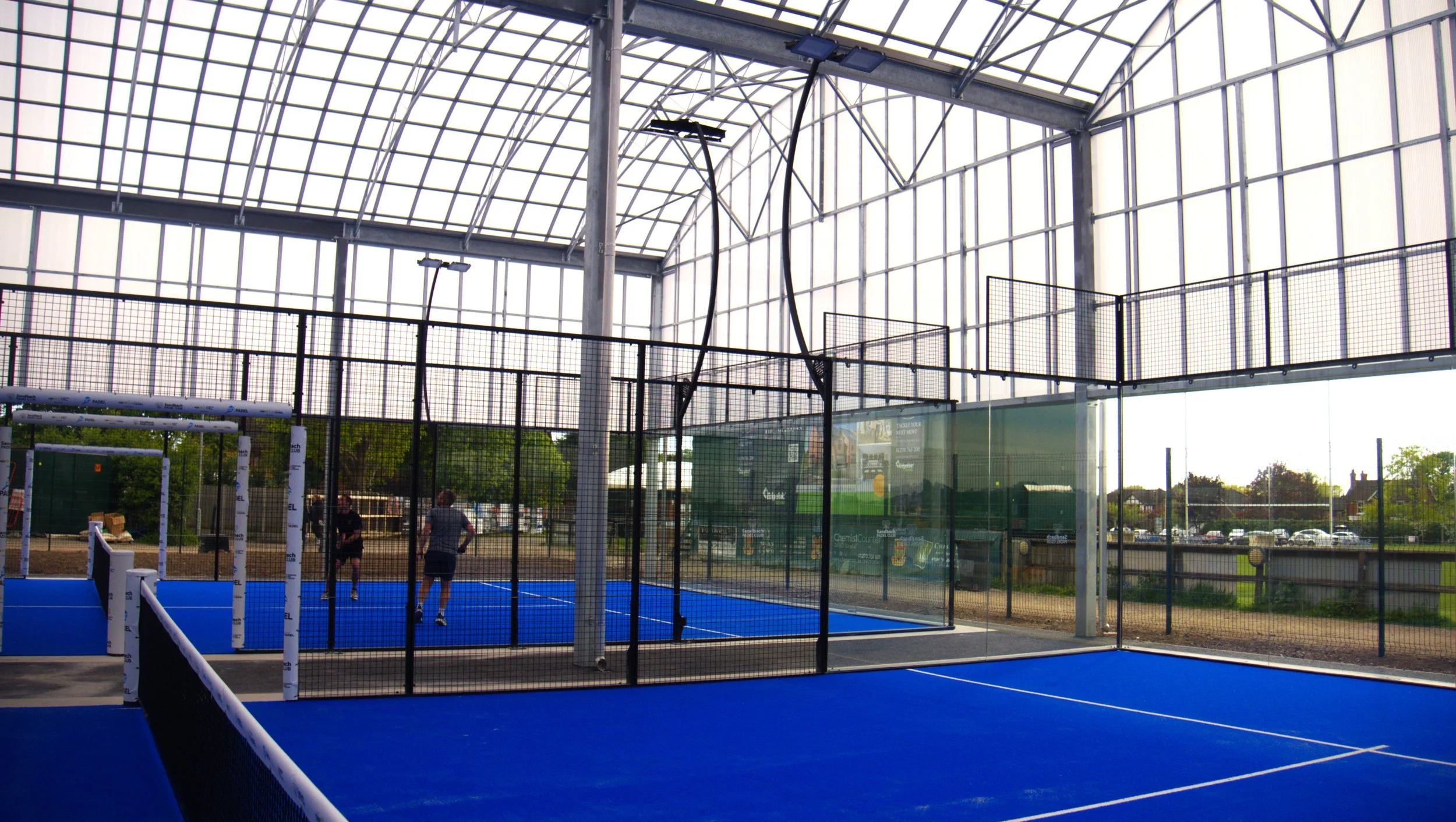 Padel facility with 3 blue padel courts, Two underneath a Canopy surrounded by a tall glass and mesh fence, with three people playing or practicing, and a large curved glass roof letting in natural light.