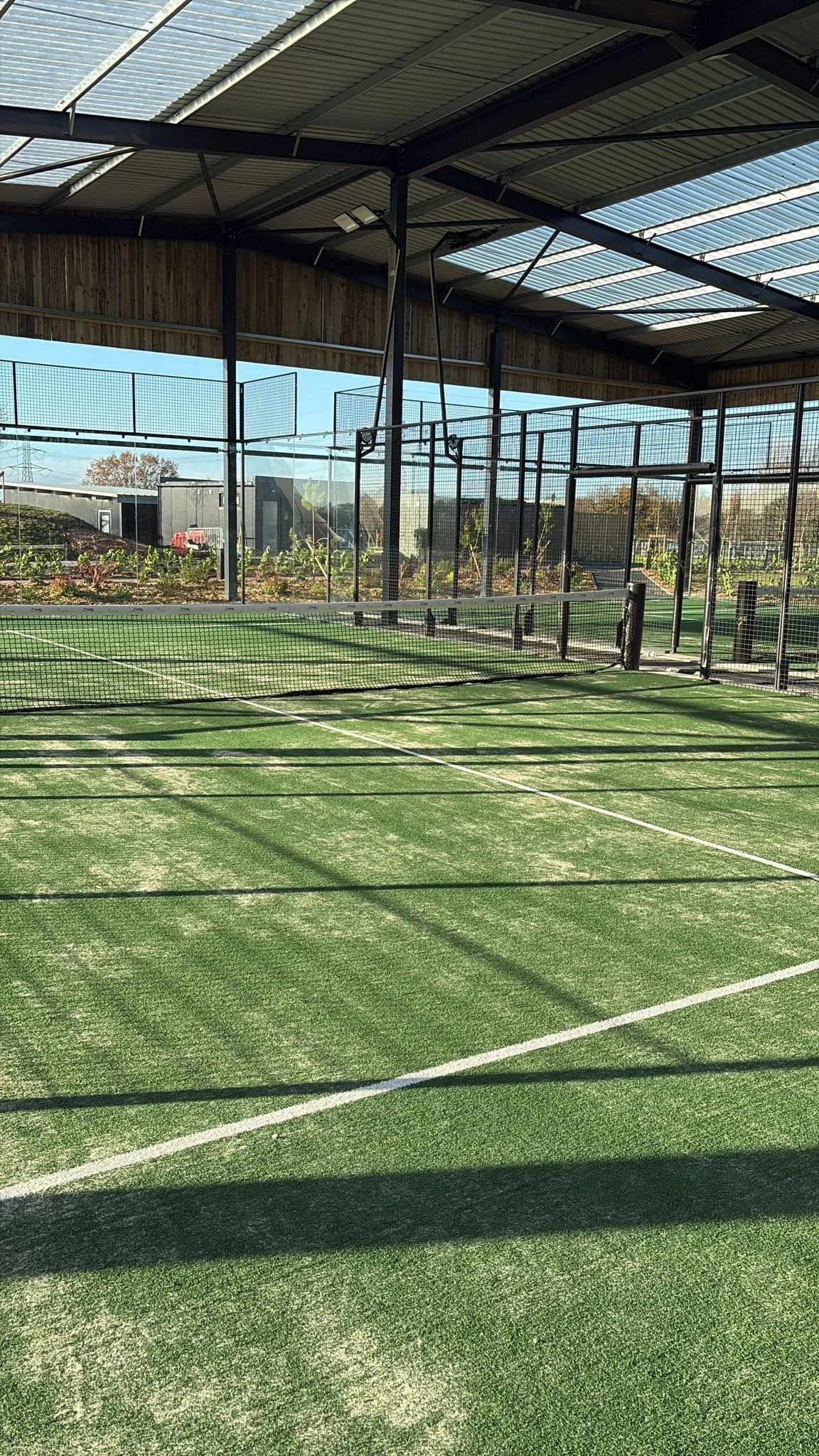 Outdoor padel tennis court with green artificial turf, surrounded by black mesh fencing and covered by a Canopy with skylights. Lake Y
