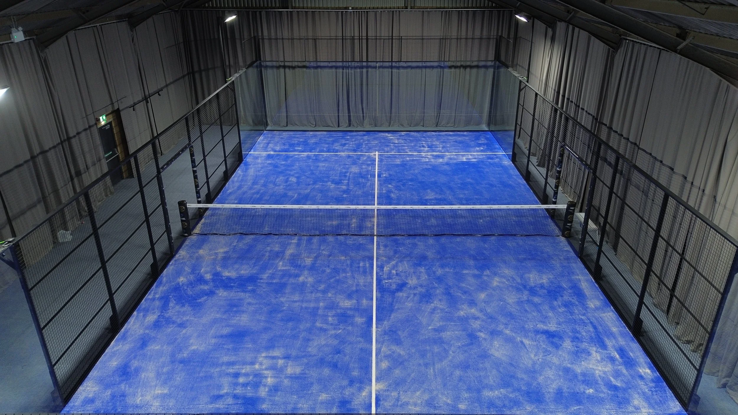 Indoor padel court with blue playing surface, surrounded by black fencing, inside a spacious warehouse with grey curtains and high ceiling.