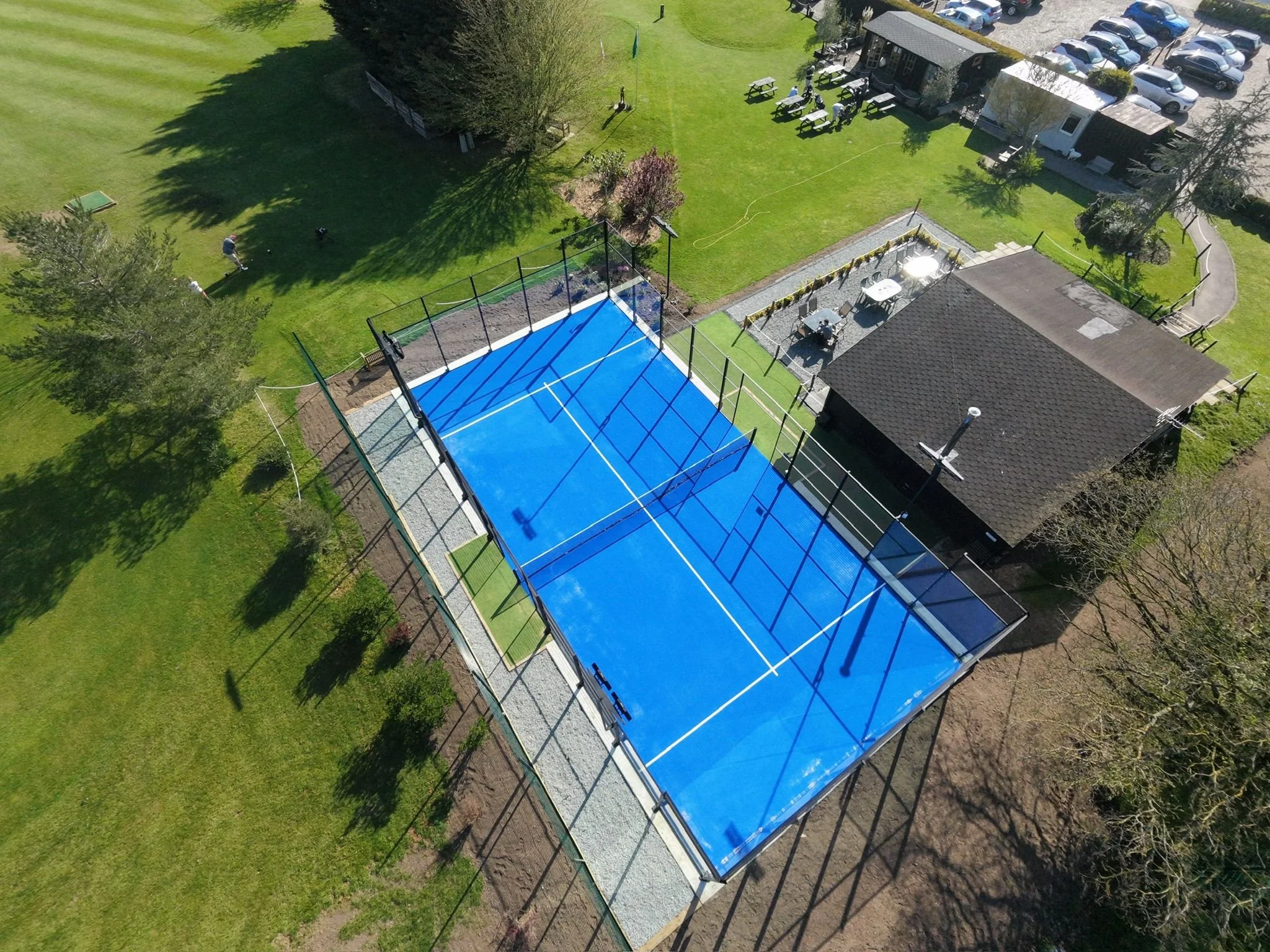 Aerial view of a park with a carom board, a Padel court, a patio with tables, and parking with numerous cars. There are trees and grassy areas, with some people and dogs visible.