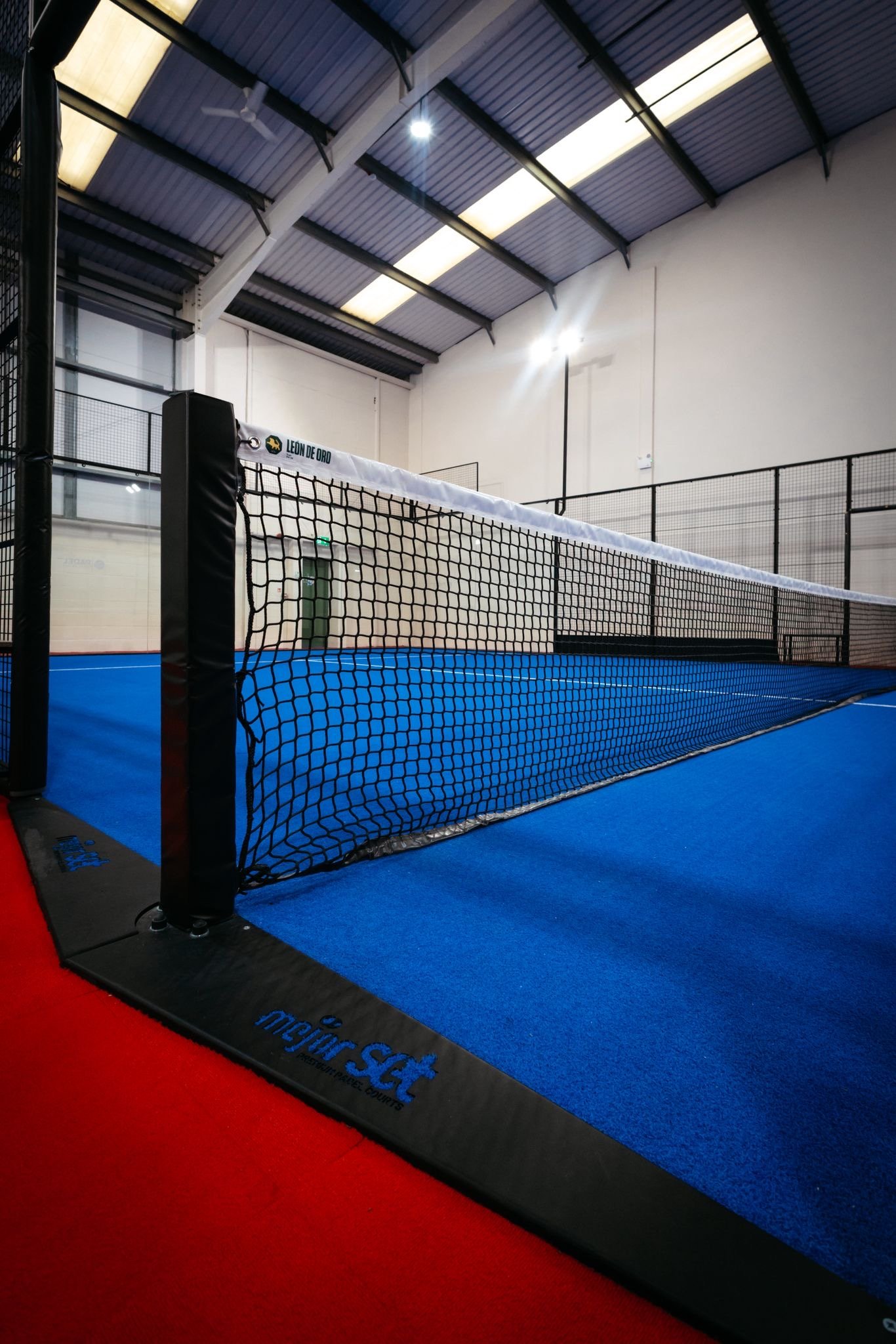 Indoor padel tennis court with blue playing surface surrounded by glass walls and black fencing UKs Best Padel