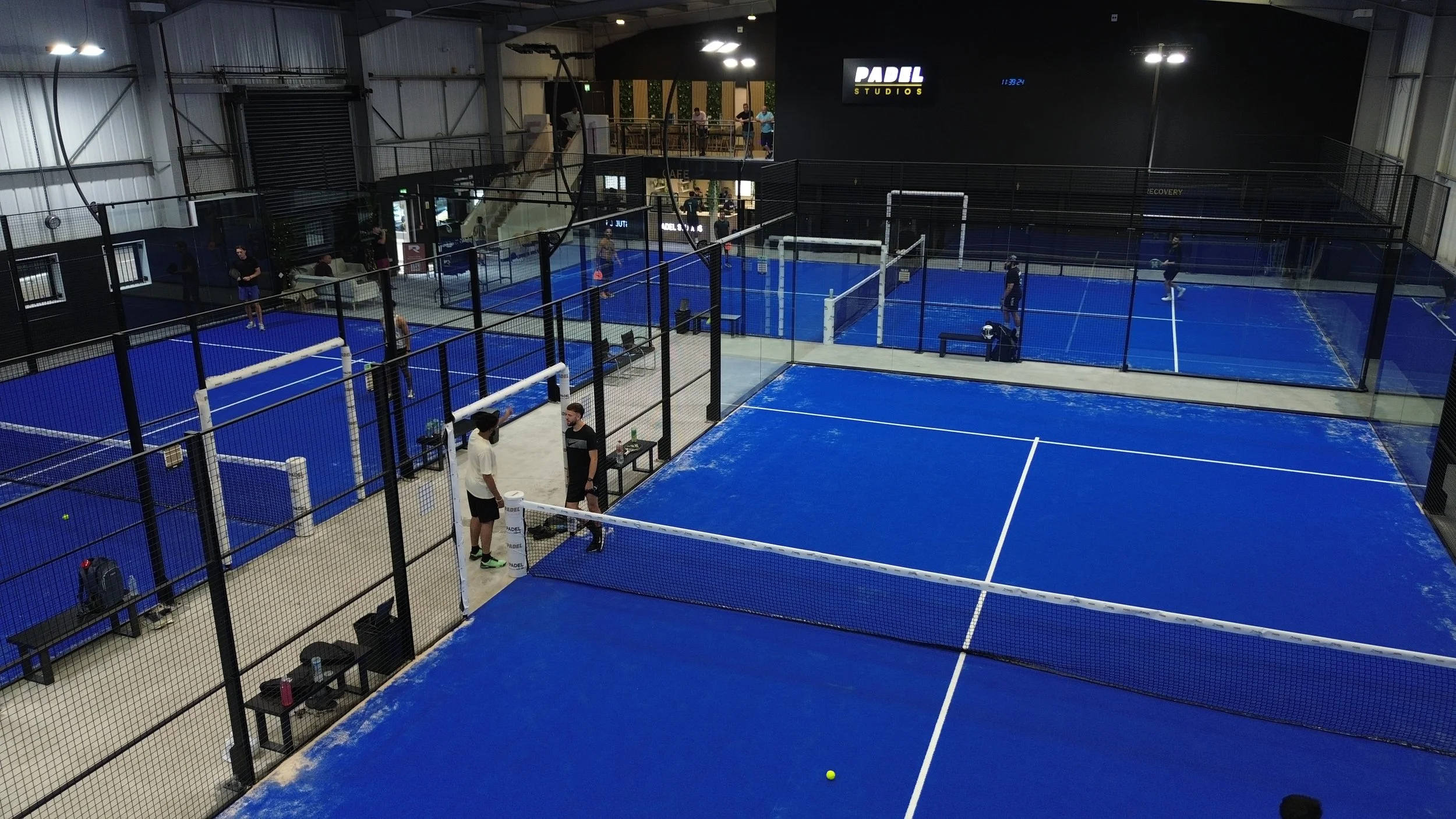 Padel Studios Coventry. An Indoor Padel Site with 3 Panoramic courts with blue turf. Players on all courts, Spectators in the background on the balcony looking over the courts. 