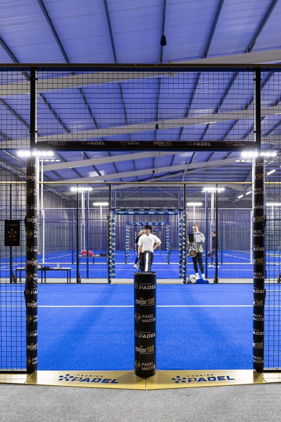 Indoor padel court with blue flooring, surrounded by metal fencing and netting, illuminated by bright overhead lights, with people standing and talking near the court entrance.