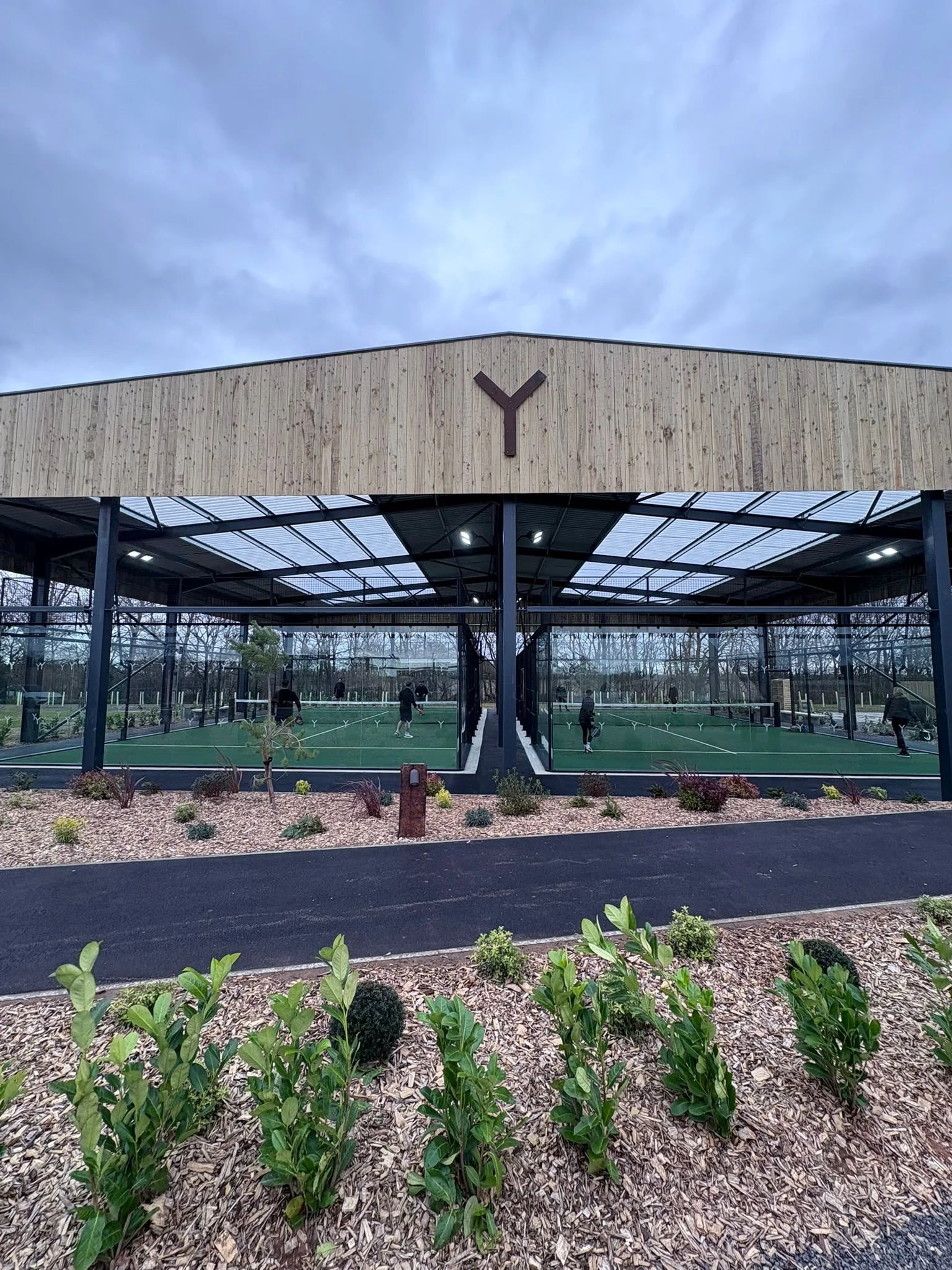 A covered Padel court with people playing, surrounded by landscaped bushes and trees, with a large wooden building in the background that has a dark Y-shaped logo on it.