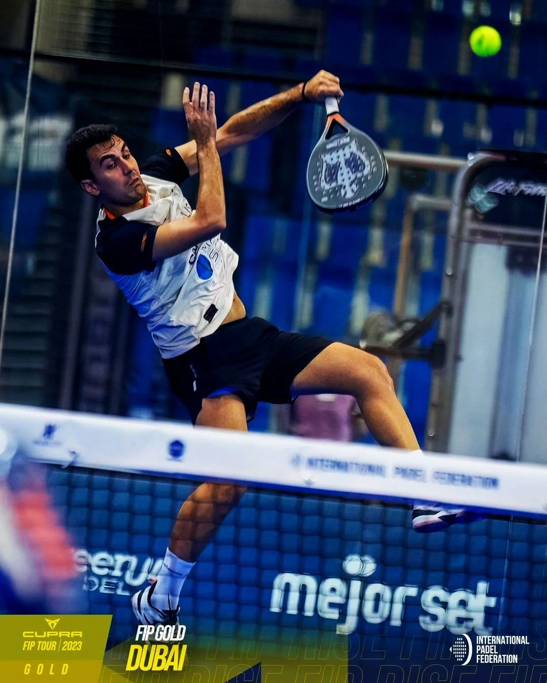 A male padel player in mid-air during a match, hitting a ball with his racket on an indoor court. He is wearing a white and black shirt and black shorts.