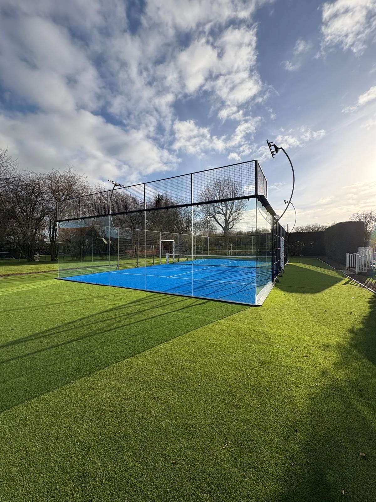 An outdoor full Panoramic Padel Court with a blue playing surface, surrounded by glass walls and fencing, under a partly cloudy sky with the sun shining on the grass court.