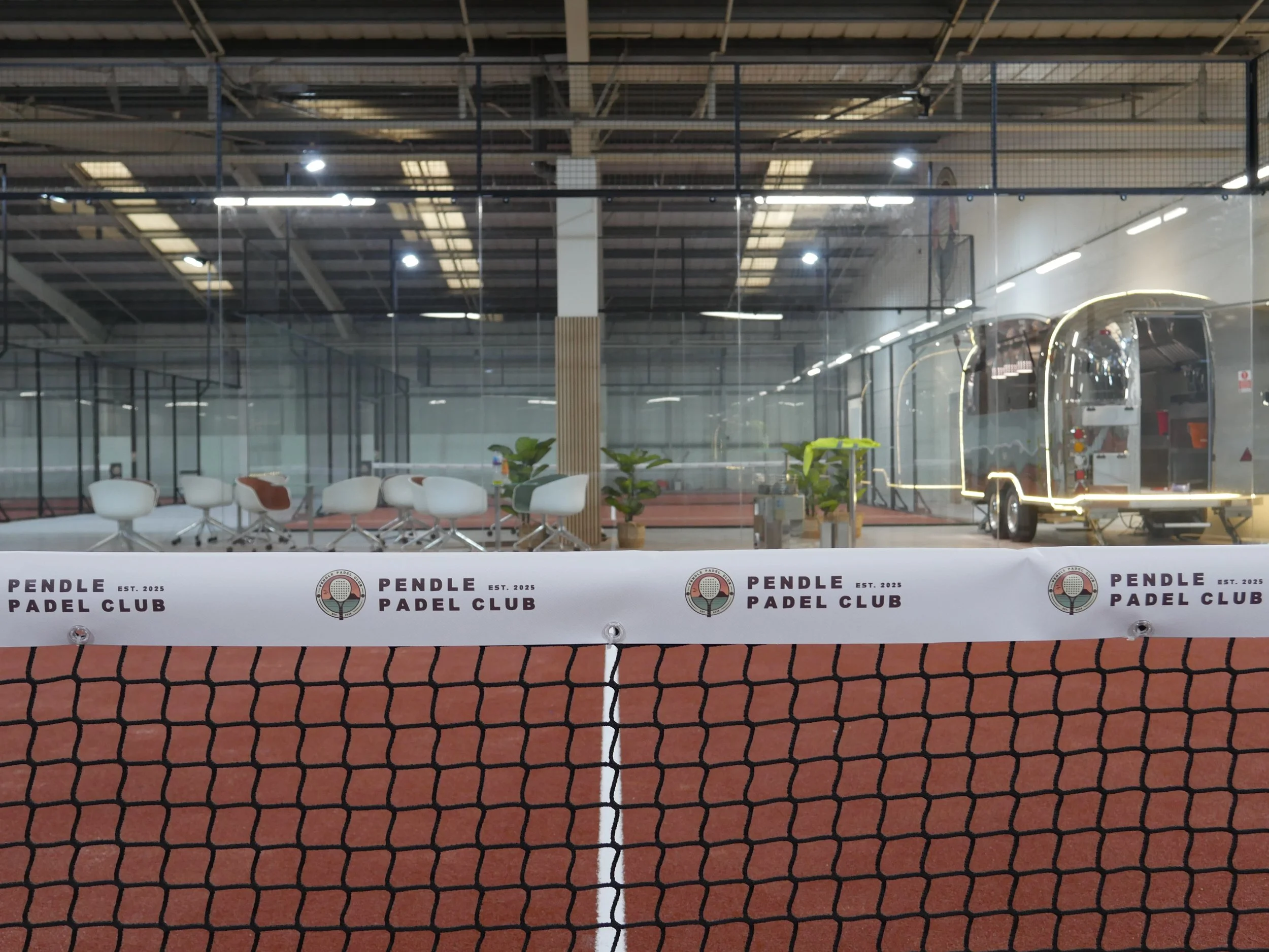 Indoor padel court with Red terracotta Turf, a net, chairs, potted plants, and a food truck or trailer, branded with the name 'Pendle Padel Club' established in 2025.