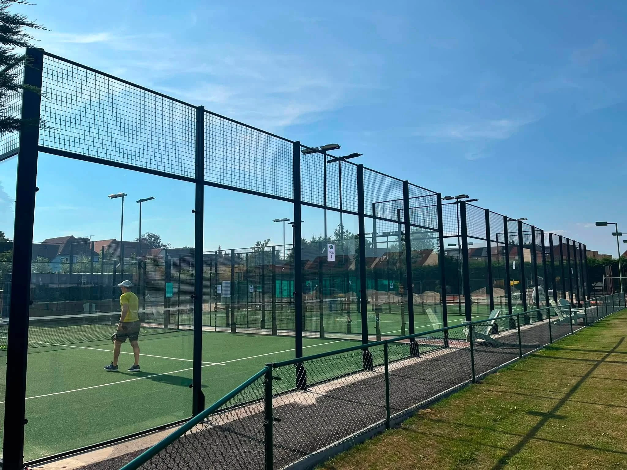 A Padel court with a high fence and pole lights, with a woman in a yellow shirt and dark shorts walking on the court under a clear blue sky.