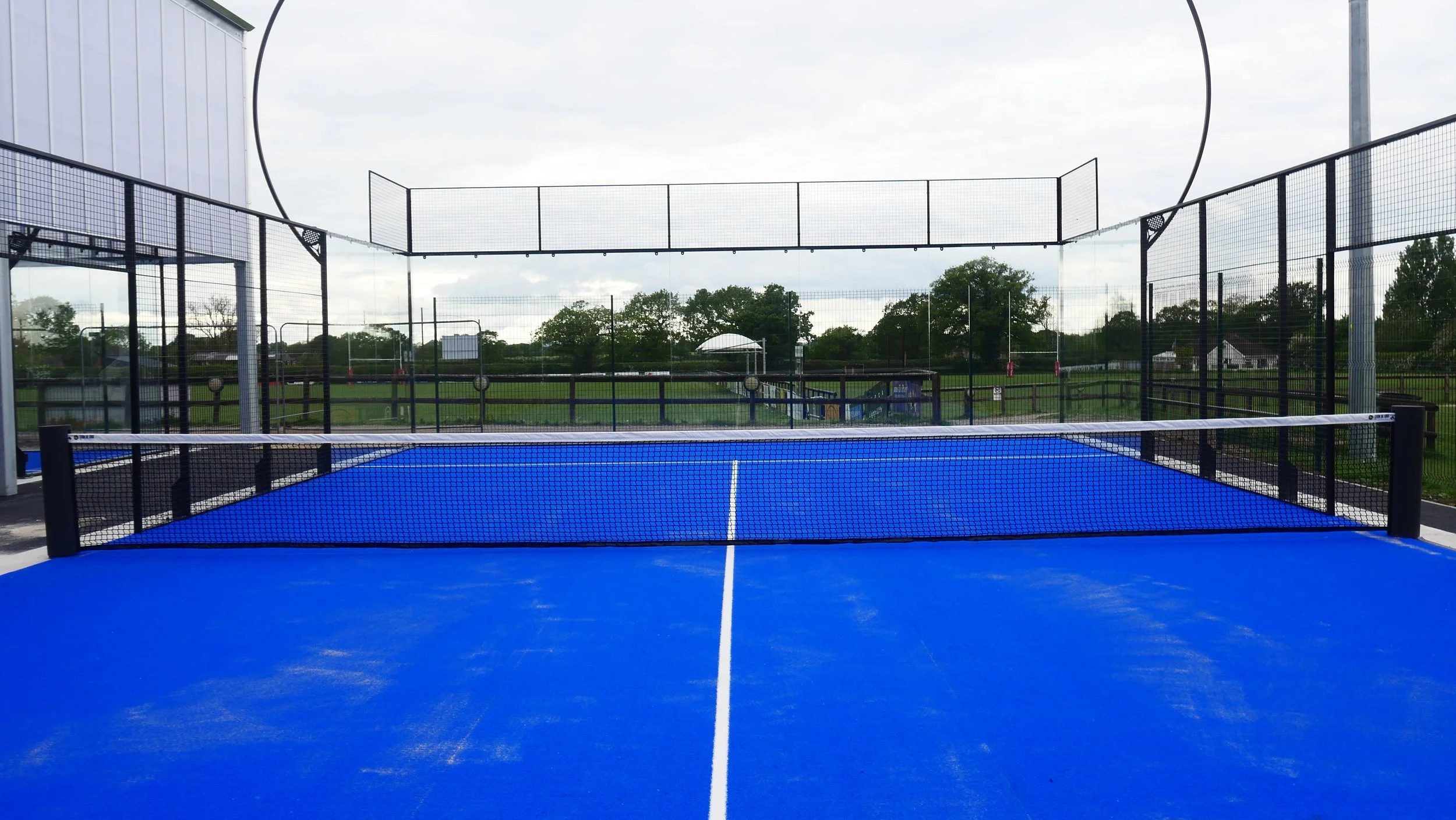 A blue Padel court with a net in the center, black fencing surrounding the court, and a cloudy sky overhead; trees and some structures are visible in the background.