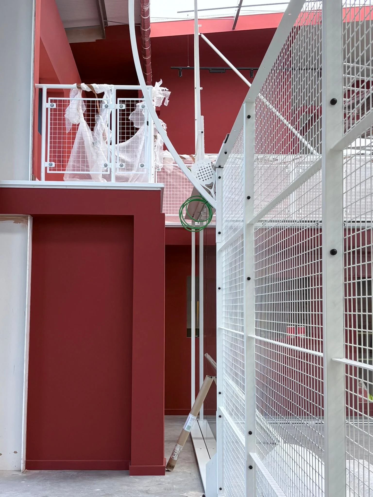 A white metal fence with a gate and a red wall of a building behind it. There are some torn white plastic coverings hanging on the fence, and a rolled-up paper tube leaning against the red wall. The floor appears to be concrete, and there are some exposed pipes and wires.