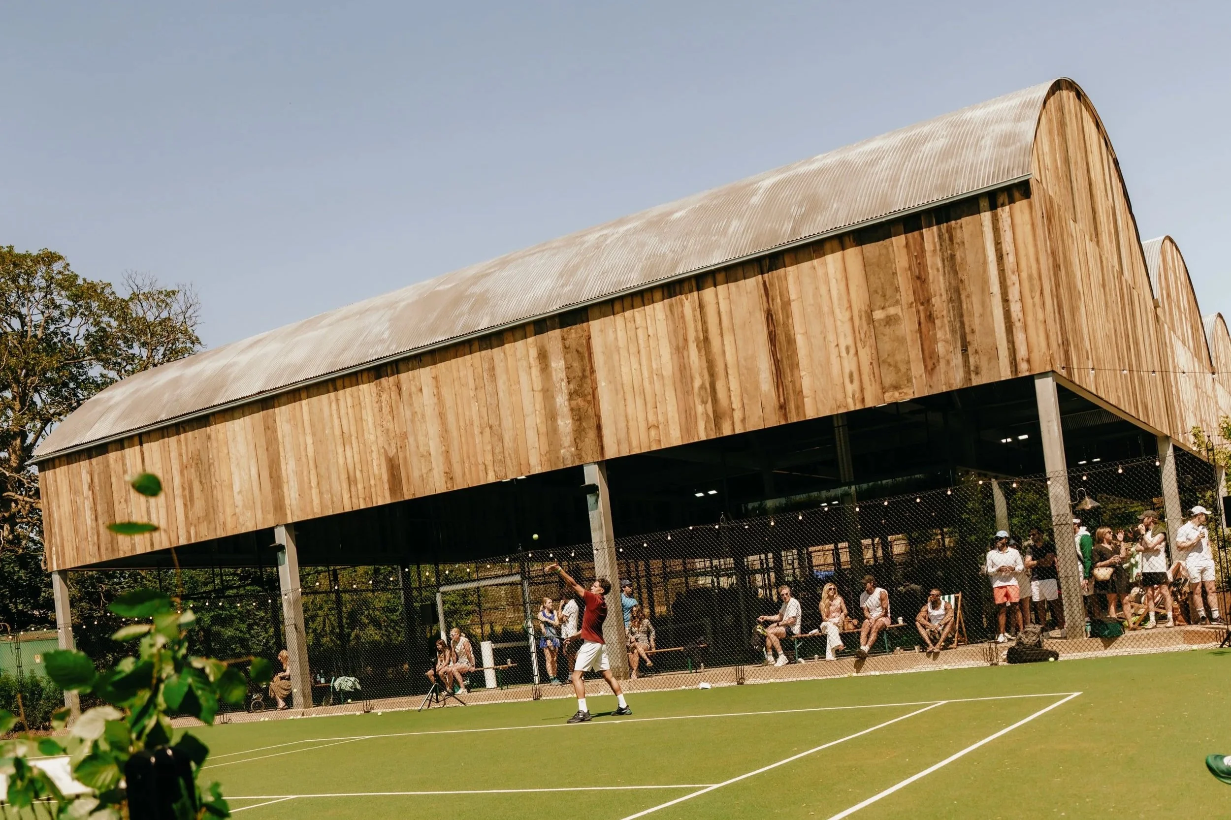 A man and a woman playing pickleball outdoors on a court with green surface. The man is wearing a black shirt, white shorts, and a white headband, holding a pickleball paddle. The woman is wearing a white tank top, white pleated skirt, white shoes, and sunglasses, holding a pickleball paddle. They are focused on the game, with lounge chairs, plants, and other people in the background.