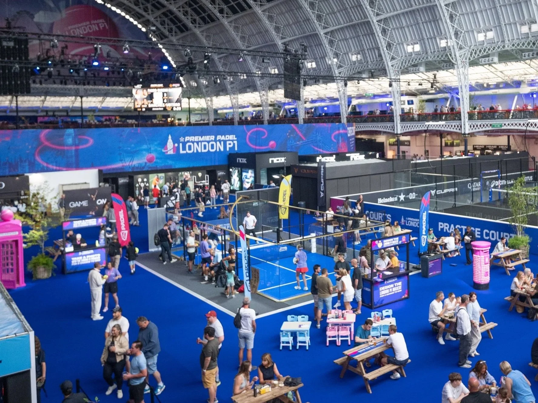 Indoor tournament venue with multiple padel courts, spectators, and banners, including advertisements for Premier Padel London P1 and Heineken, with groups of people walking and standing around.