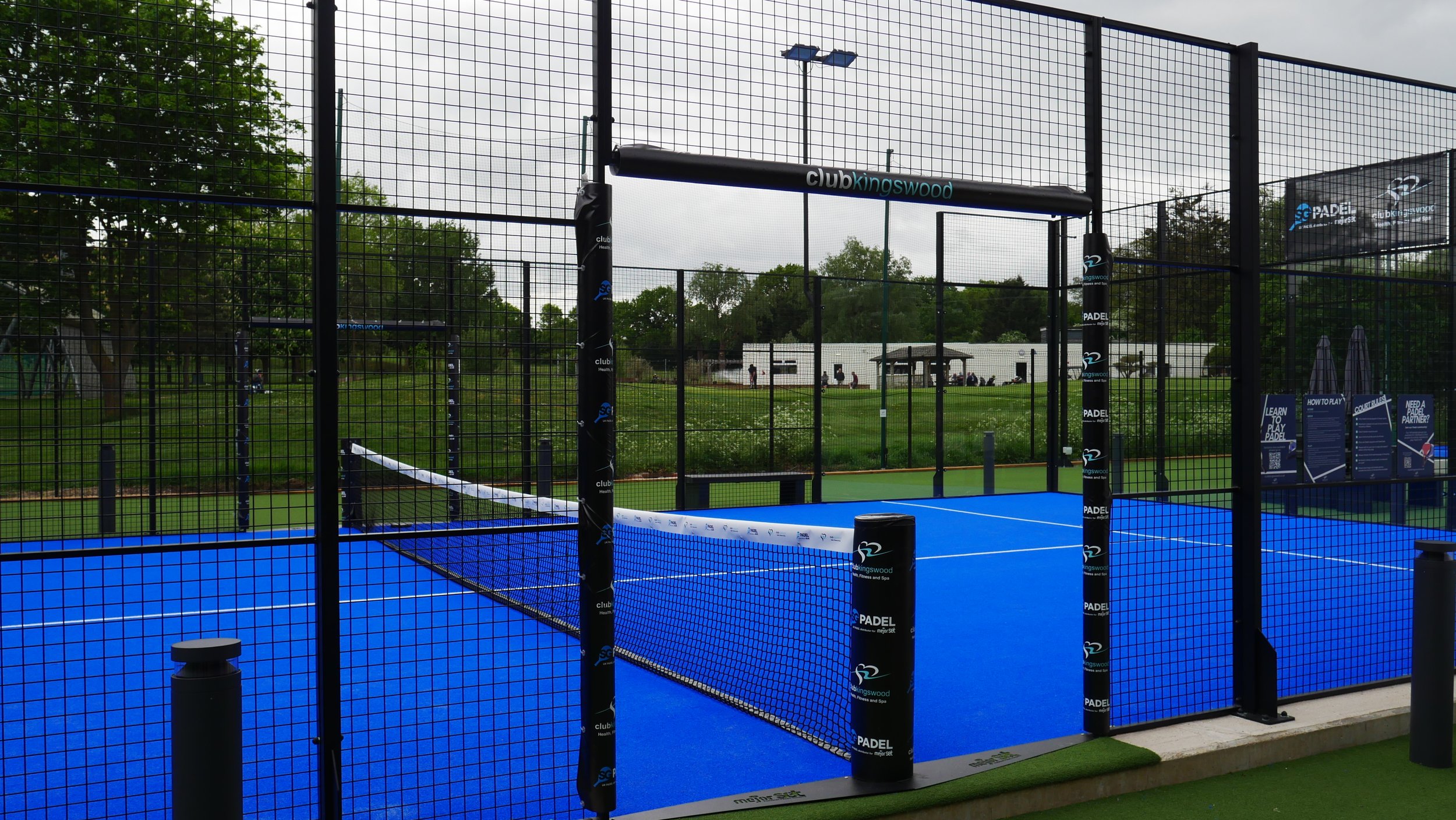 An outdoor padel court with a bright blue playing surface, surrounded by a black metal fence, with trees and a grassy area in the background.