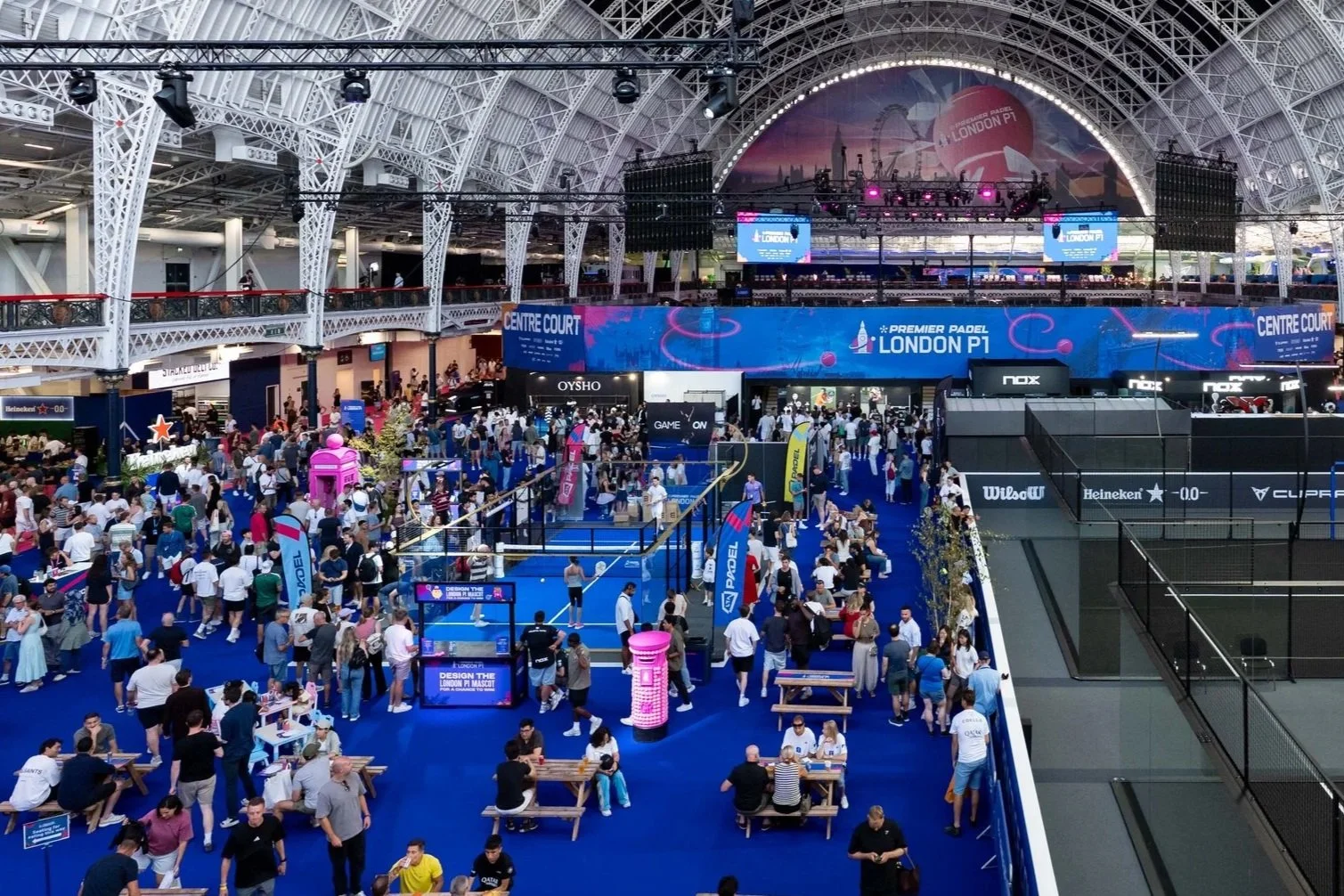 Indoor tournament venue with crowds, featuring a large blue banner reading 'Premier Padel London P1', multiple tennis or padel courts, seating areas, and overhead screens displaying event information.