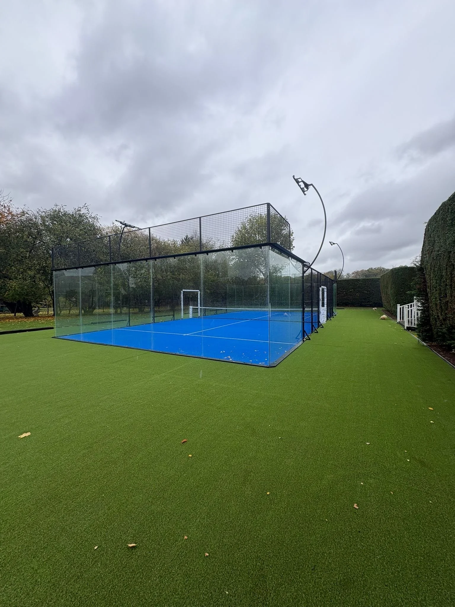 An outdoor padel tennis court with blue surface, Surrounded by green turf.