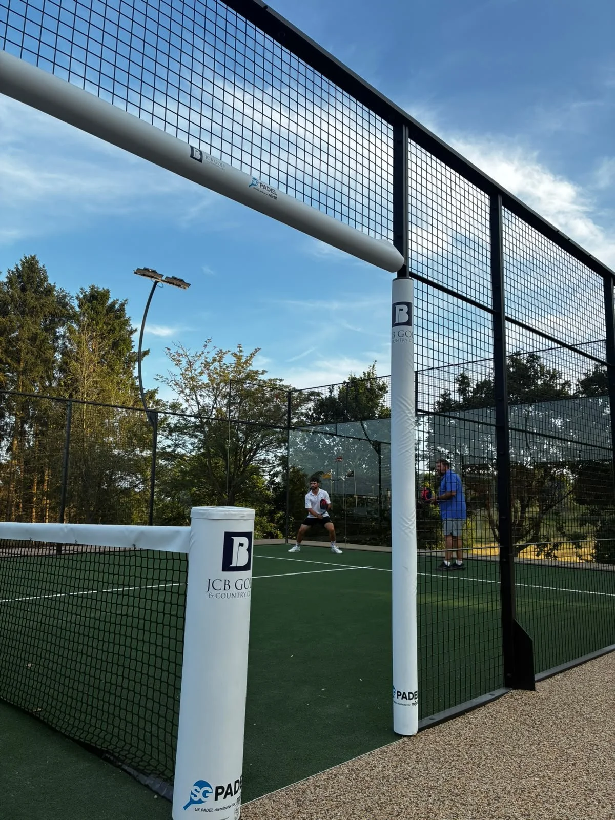 Two men playing padel tennis on an outdoor court with a net, surrounded by a tall black fence and trees, under a clear blue sky.
