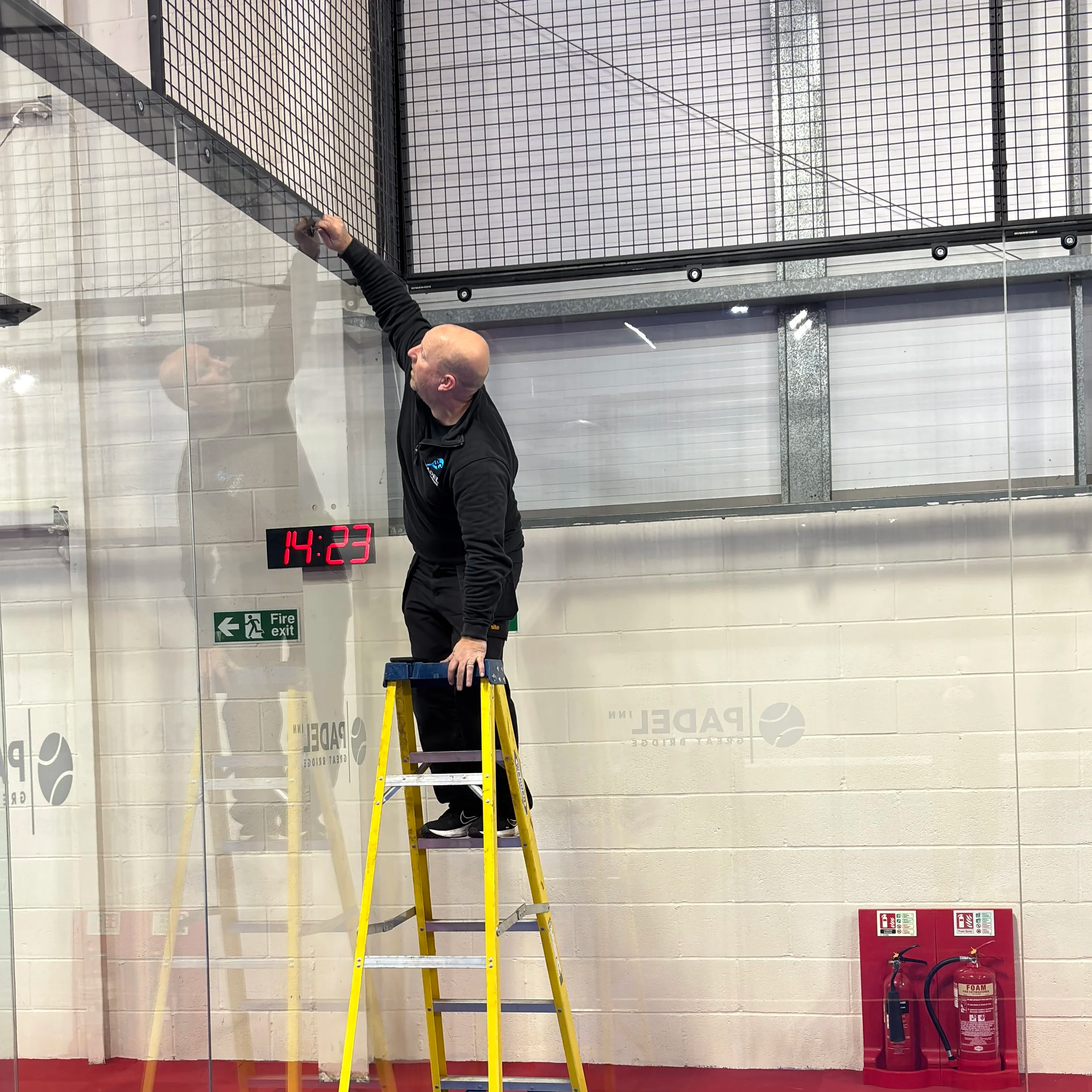 A Man cleaning the glass and tightening screws on a Padel Court, wearing a Jacket that says SG Padel