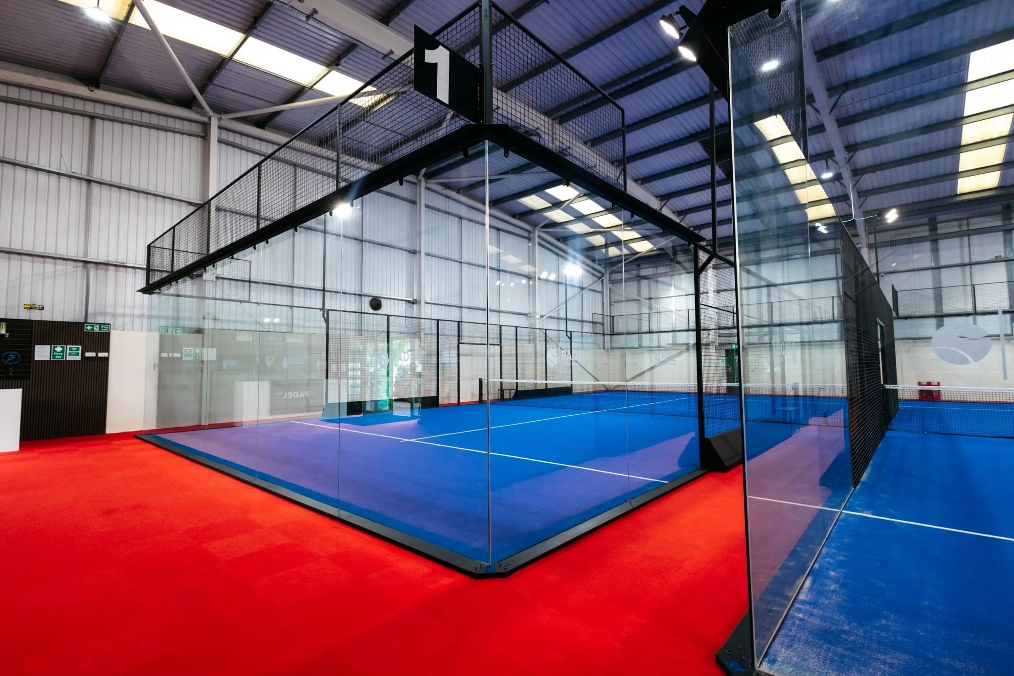 Indoor padel tennis court with blue playing surface surrounded by glass walls and black fencing UKs Best Padel