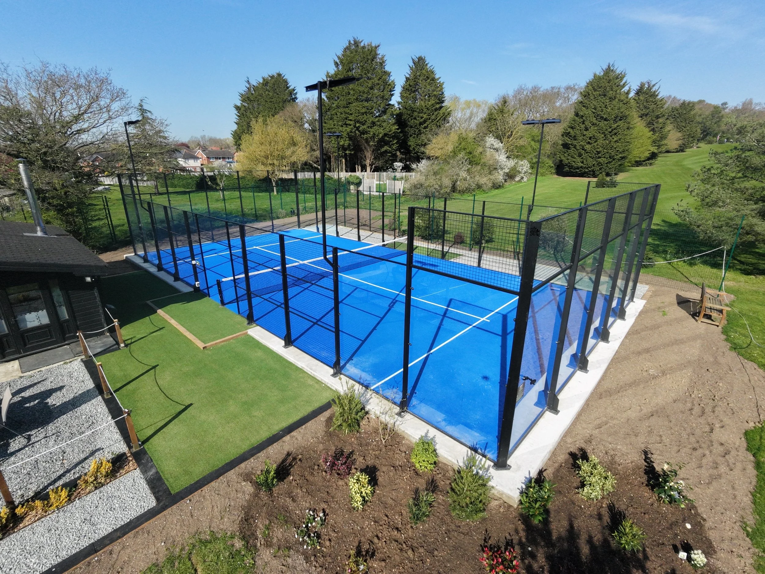 A Golf course with a blue Padel court, surrounded by a black fence and tall trees, with a lawn and residential houses in the background.